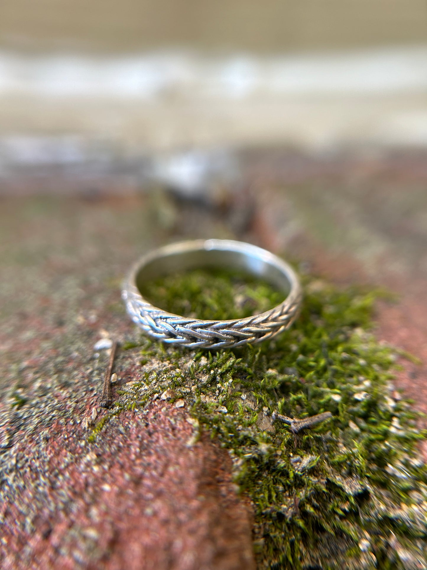 Close-up of a Canyon Jewelry sterling silver braided chain ring resting on green moss, with a blurred background emphasizing the detailed texture of the ring and moss surface.