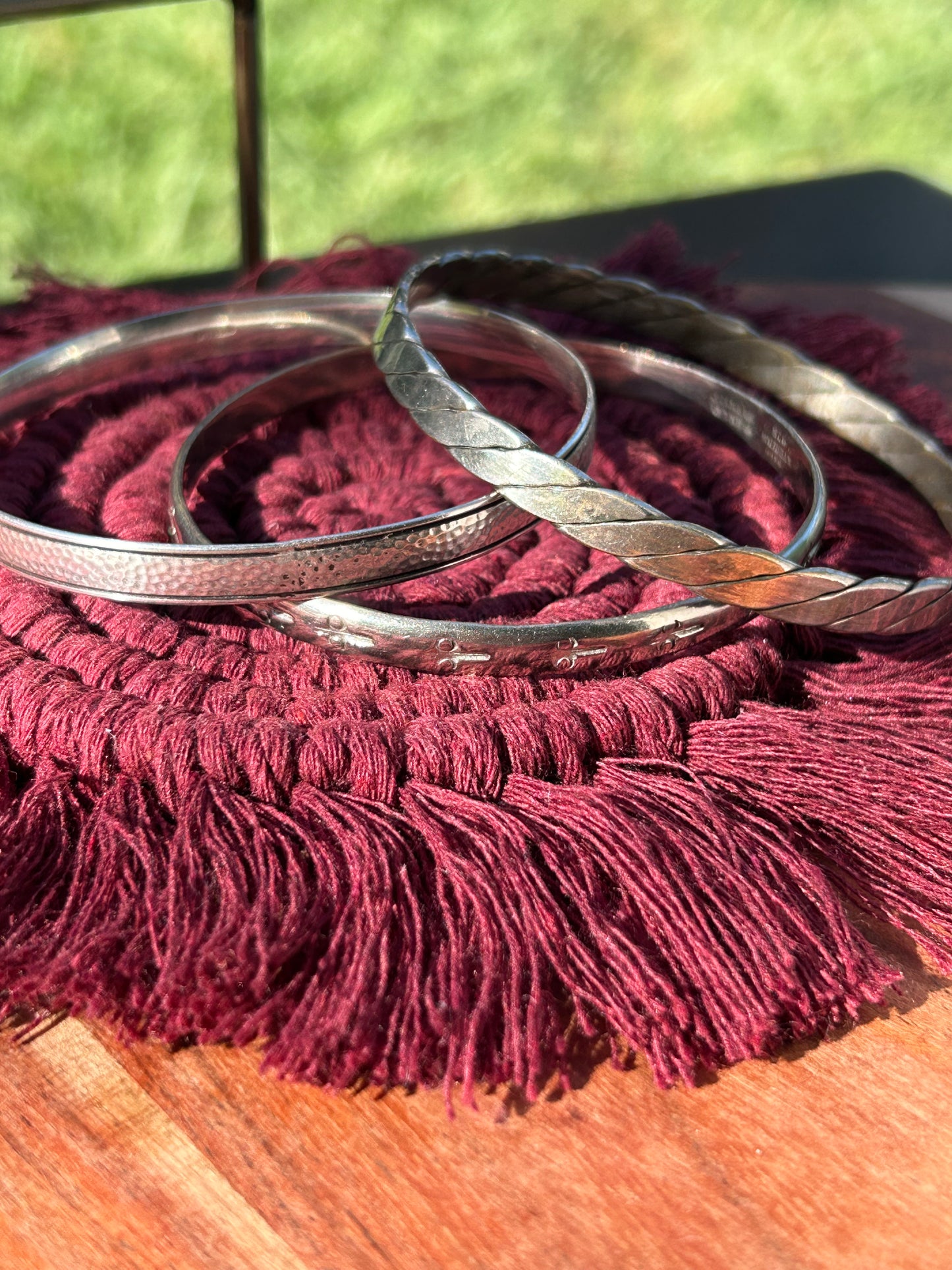 Close-up of a Canyon Jewelry Pathway Bangle beside a white vase, resting on a burgundy tasseled cloth under sunlight, highlighting textured details on the bracelet and contrasting shadows.