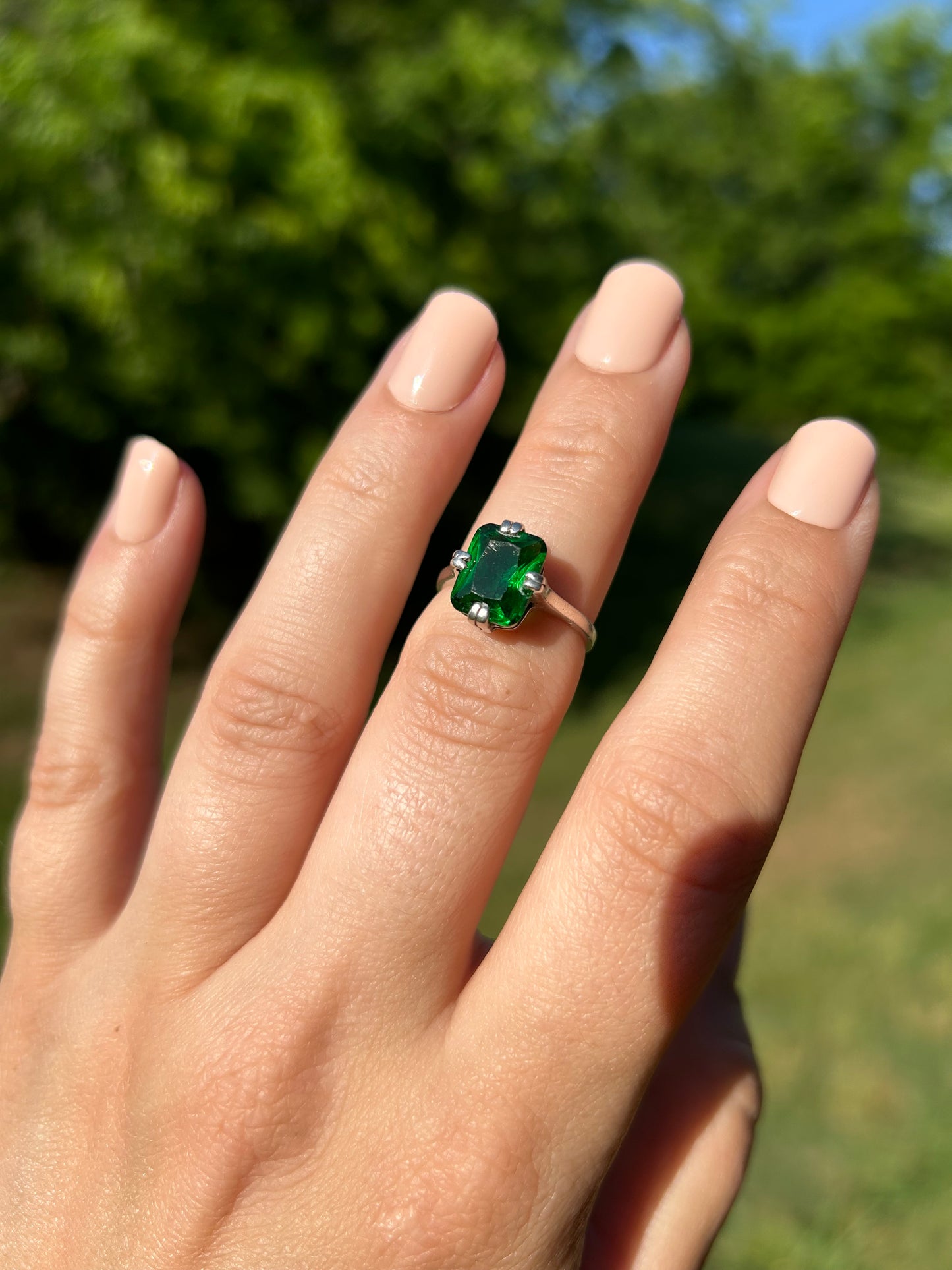 A close-up of a hand holding an Enchanted Forest Ring from Canyon Jewelry with a large, green gemstone, set against a softly blurred green grass background, capturing a vintage style.