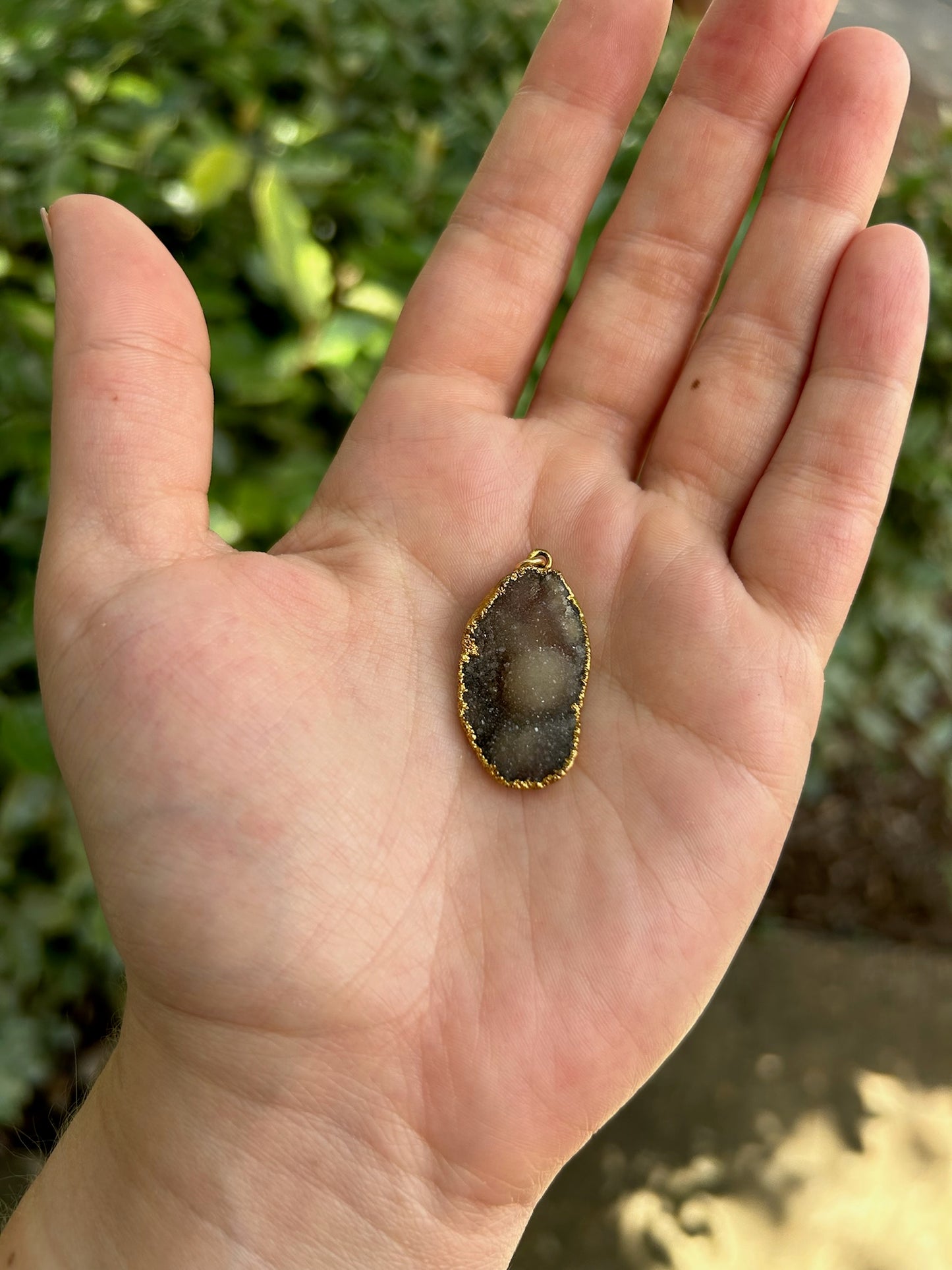 Close-up of a translucent, oval Druzy Agate pendant with a sparkling, crystalline interior and a sterling silver rim, resting on a moss-covered rock, with a blurred background. (Brand Name: Canyon Jewelry)