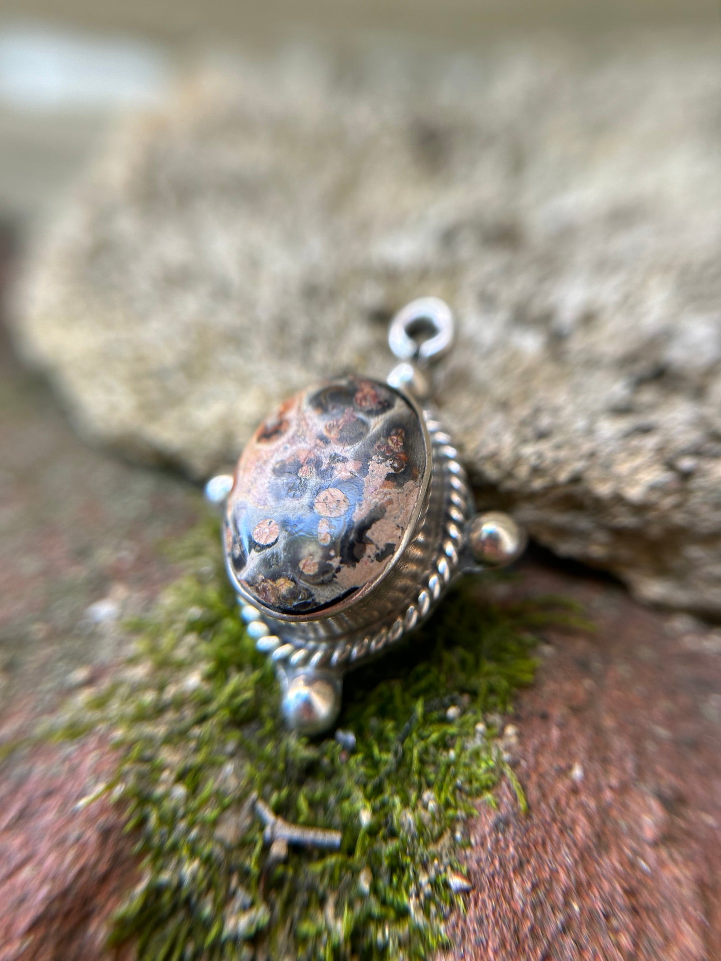 Close-up of an intricately designed Canyon Jewelry Volcano Pendant with a speckled oval stone, set in a sterling silver frame, resting on a mossy surface. The background is blurred with a focus on the detailed craftsmanship.