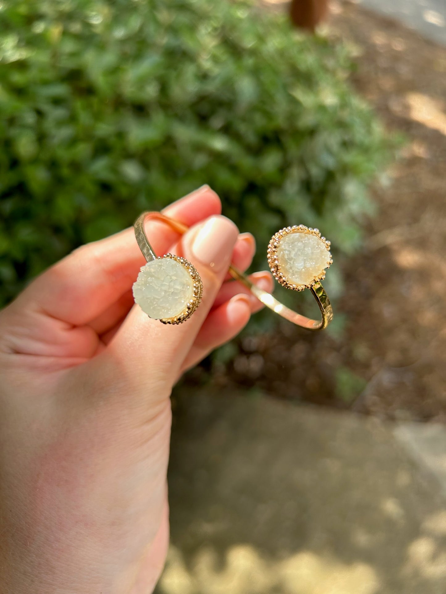 A close-up of a hand holding two Divergence Bracelets from Canyon Jewelry, with large, white, sparkly circular pendants, focuses against a softly blurred greenery background. These statement jewelry pieces capture attention effortlessly.