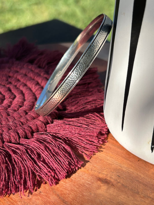 Close-up of a Canyon Jewelry Pathway Bangle beside a white vase, resting on a burgundy tasseled cloth under sunlight, highlighting textured details on the bracelet and contrasting shadows.