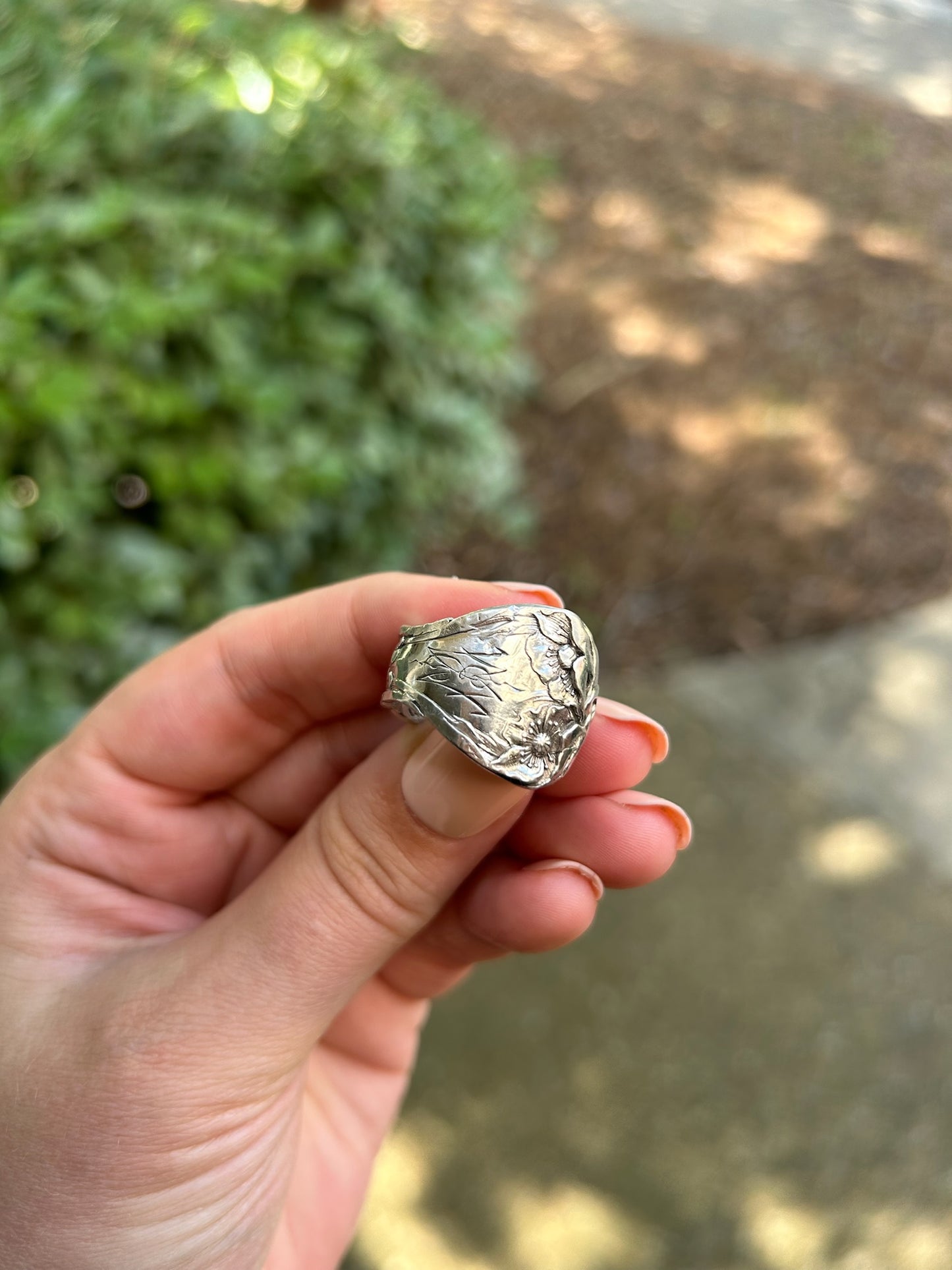 A close-up of a Canyon Jewelry 1913 Spoon Ring resting on a green mossy surface, depicting floral designs with prominent details against a blurred background.