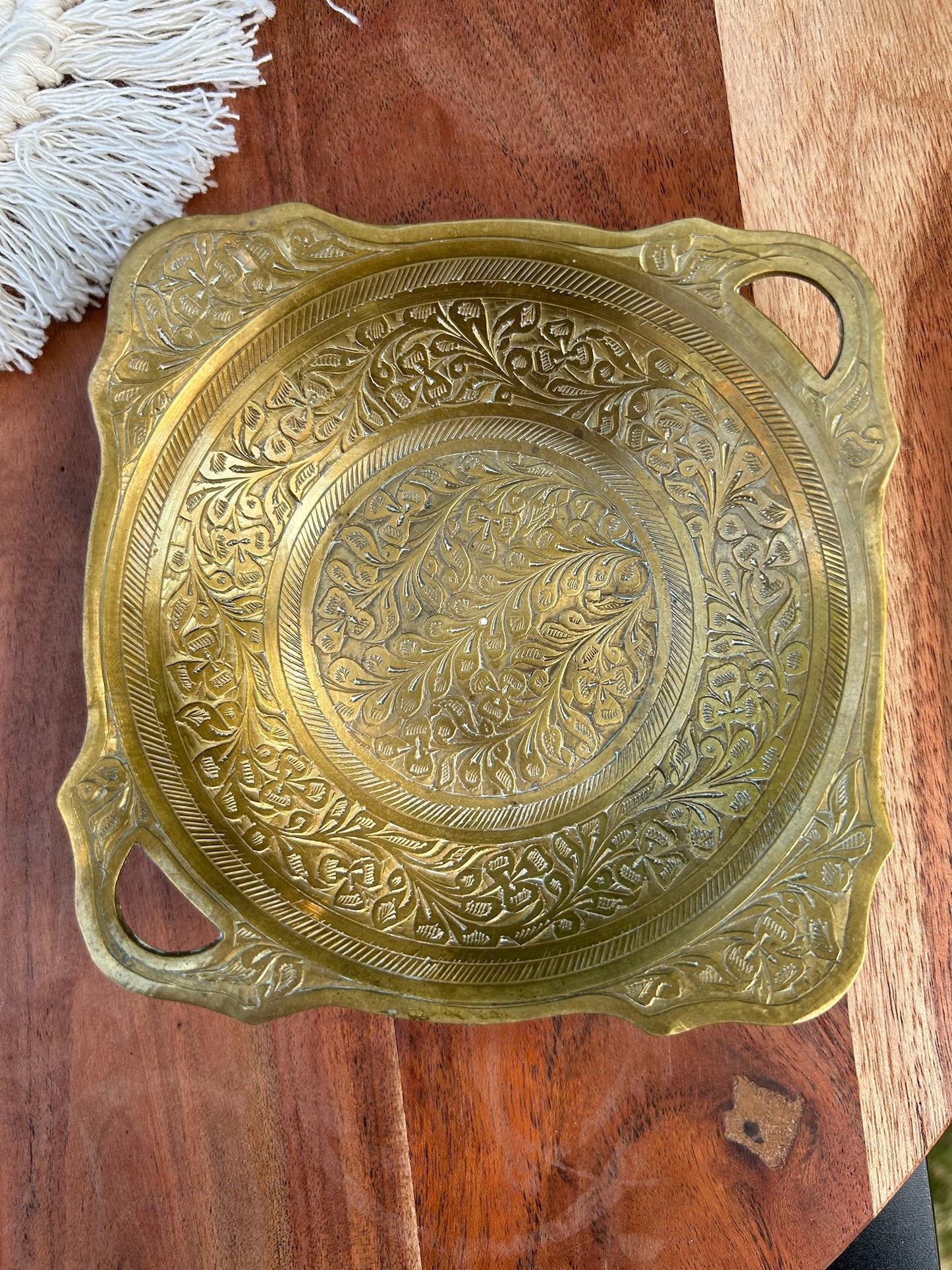 An intricately designed Canyon Jewelry Brass Trinket Tray with boho jewelry patterns placed on a wooden surface outdoors, partially shaded by sunlight.