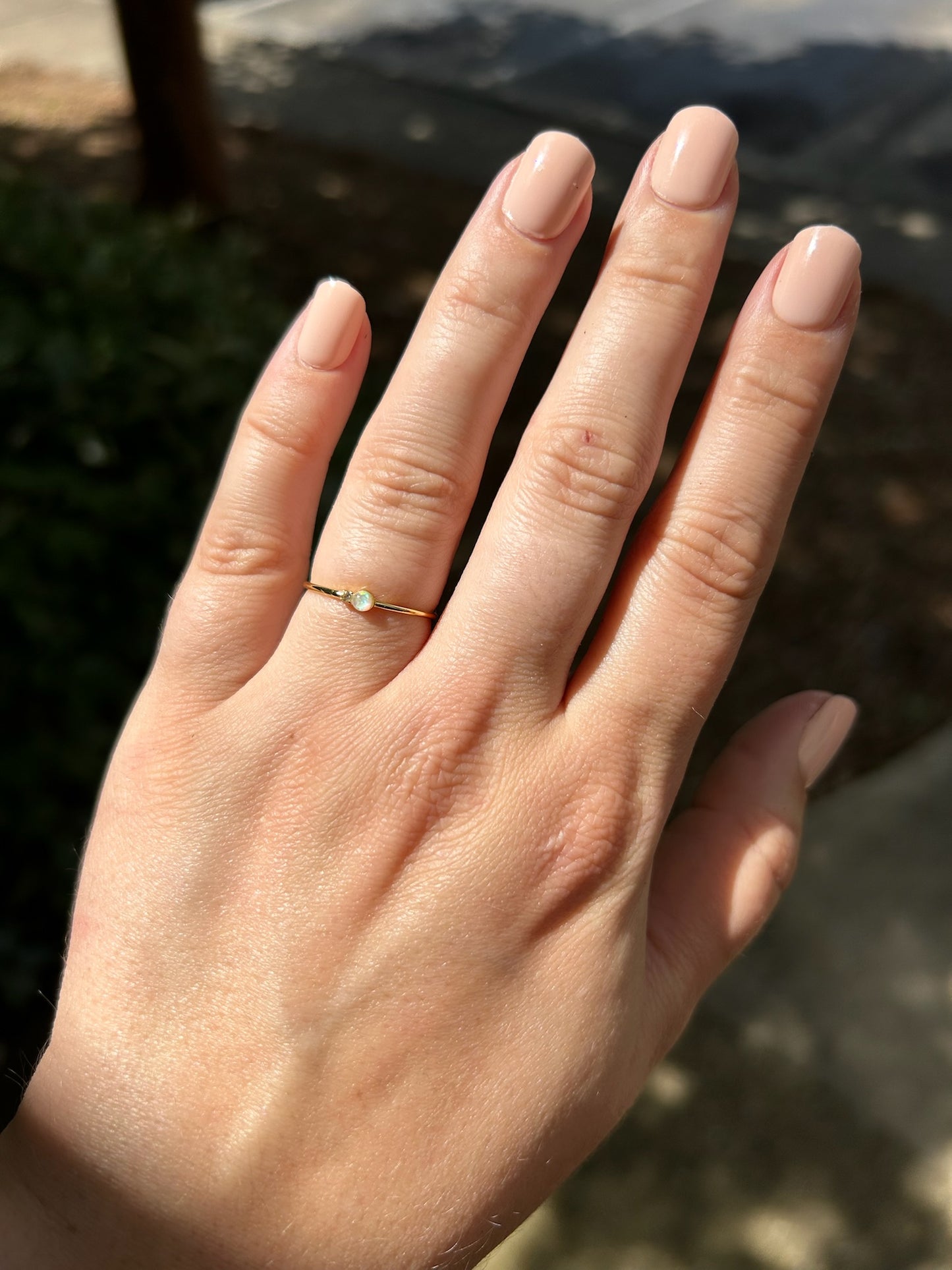 Close-up of a delicate Selene Ring by Canyon Jewelry, with a small opal set in it, resting on a textured surface covered with moss and tiny red plants. The background is softly blurred, evoking a boho vibe.