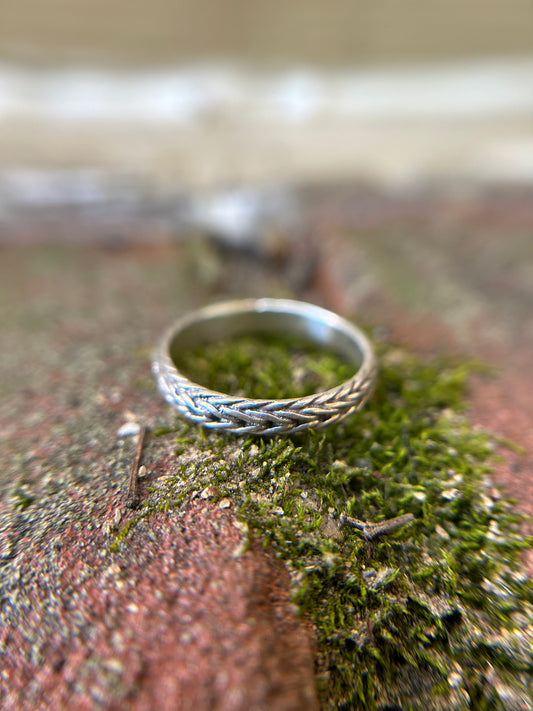 Close-up of a Canyon Jewelry sterling silver braided chain ring resting on green moss, with a blurred background emphasizing the detailed texture of the ring and moss surface.