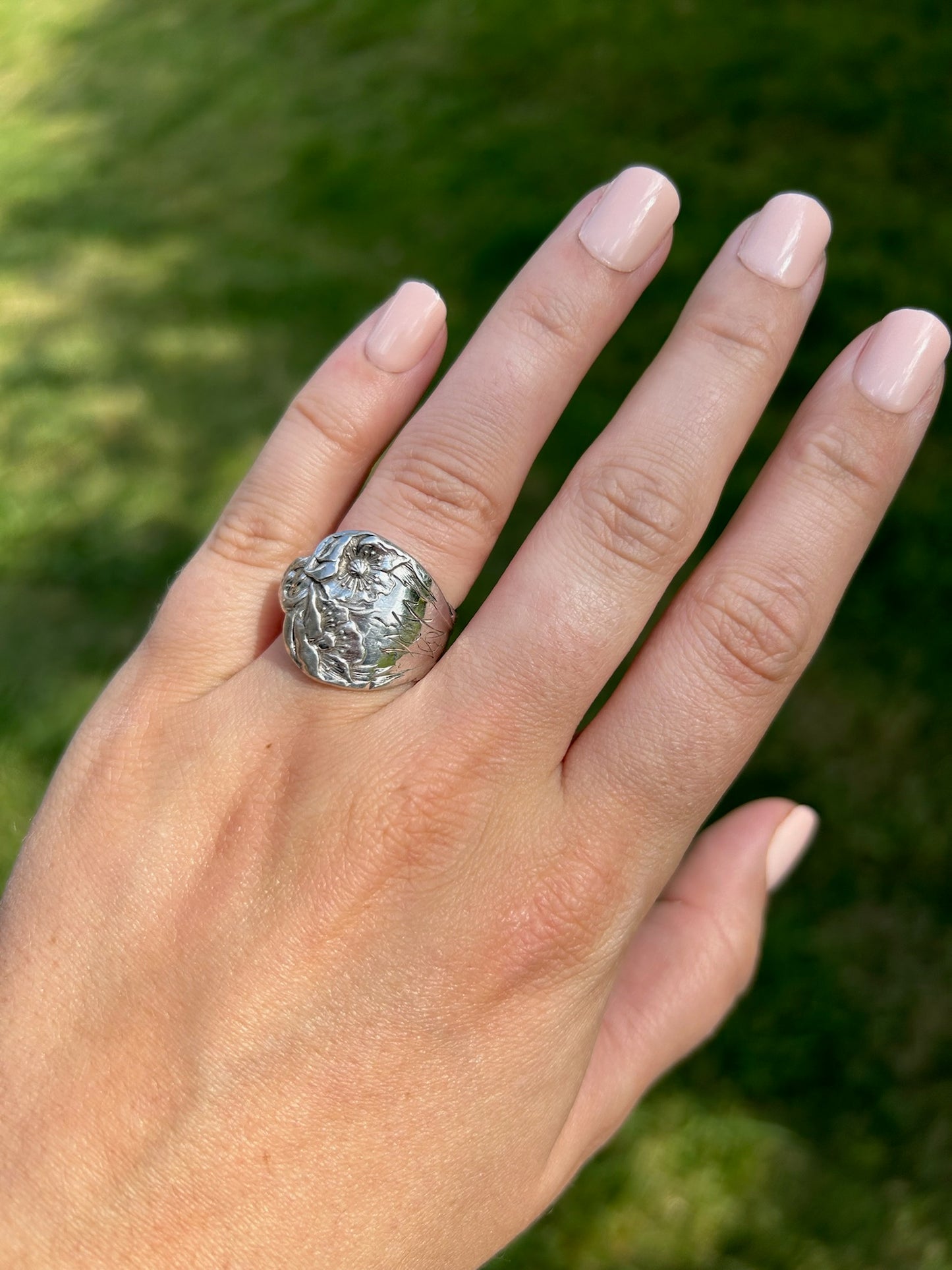 A close-up of a Canyon Jewelry 1913 Spoon Ring resting on a green mossy surface, depicting floral designs with prominent details against a blurred background.