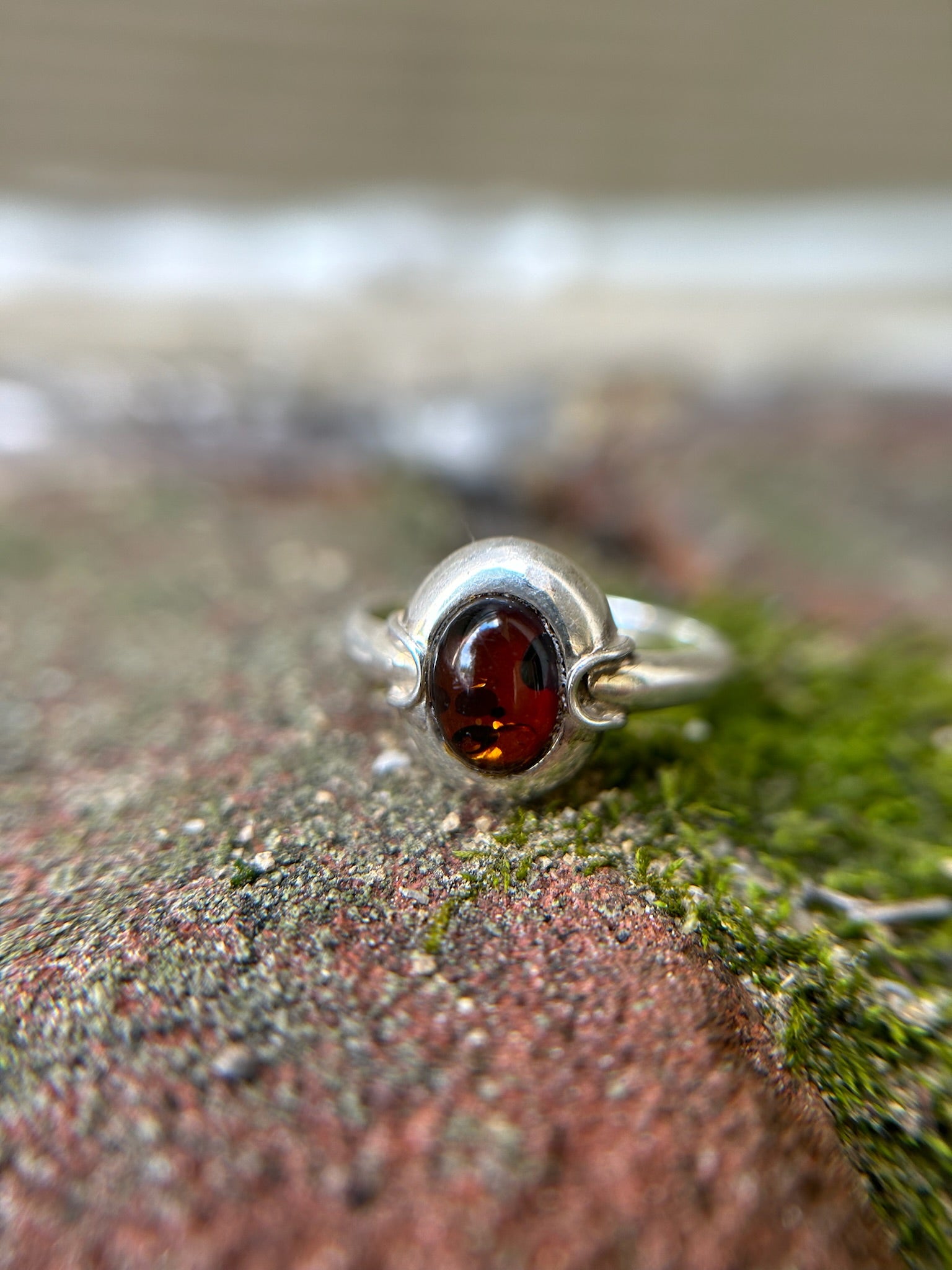 Close-up image of a vintage Ladybug Ring from Canyon Jewelry with an amber gemstone, set against a background of moss-covered stone. The focus is sharp on the gemstone, highlighting its rich, deep color.