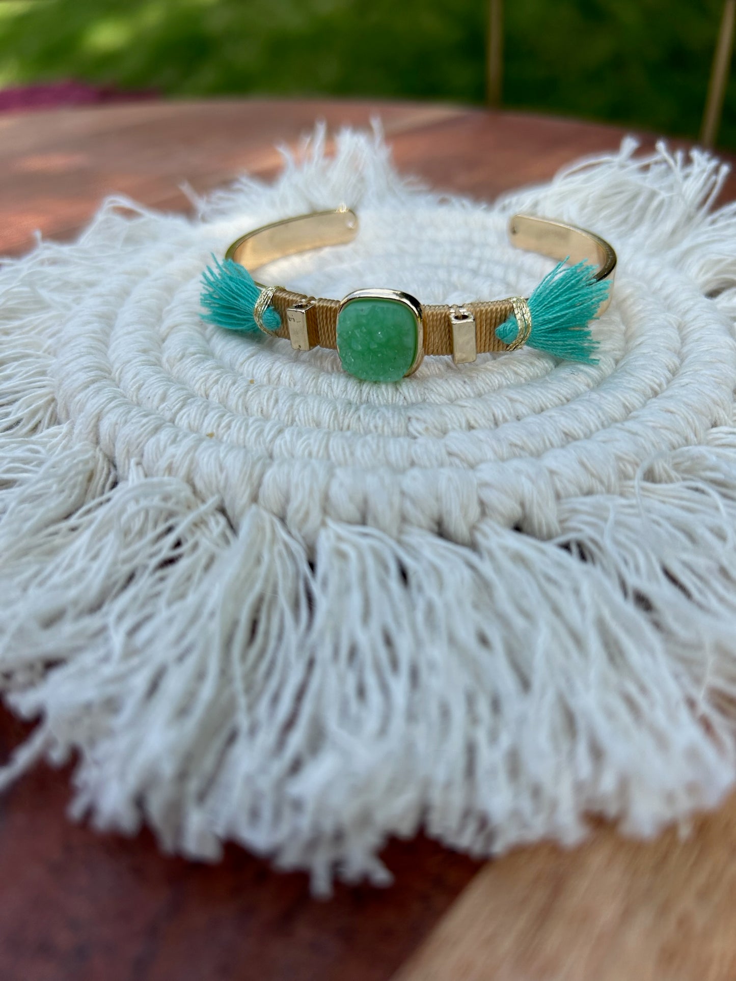 A person's wrist adorned with a stylish Painted Pony Bracelet from Canyon Jewelry featuring a large green oval centerpiece and turquoise tassel details, set against a soft-focus outdoor background. This piece exemplifies statement jewelry.