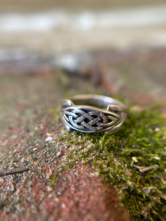 Close-up of a silver Canyon Jewelry boho celtic knot ring resting on a mossy surface, with a soft-focus background that includes indistinct natural elements.