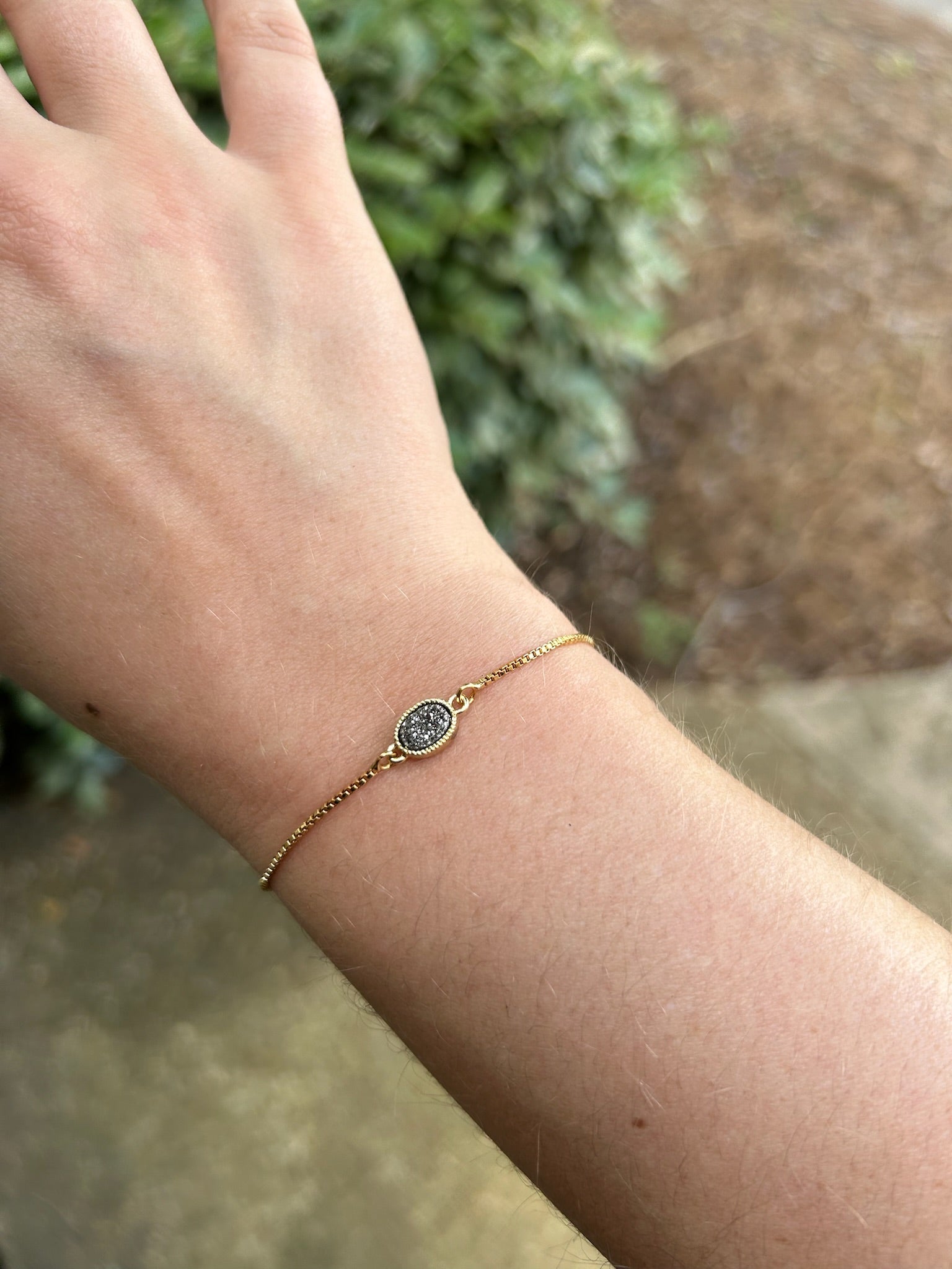 A Pedal to the Metal Bracelet with a delicate chain and a small, circular, glittering pendant lies on a wooden surface next to a burgundy fabric with fringe in western fashion.