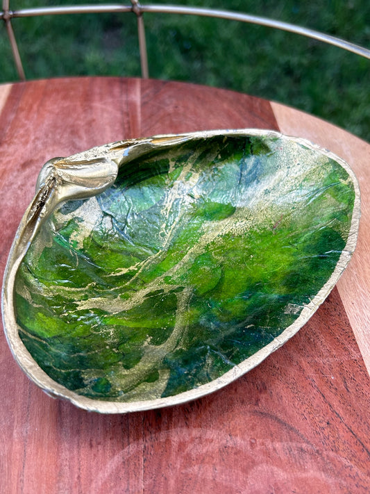 A decorative ceramic dish shaped and painted to resemble a green leaf with vein details, displayed on a sterling silver surface. The Canyon Jewelry Green Shell Trinket Tray has subtle golden accents along the edge and handle.