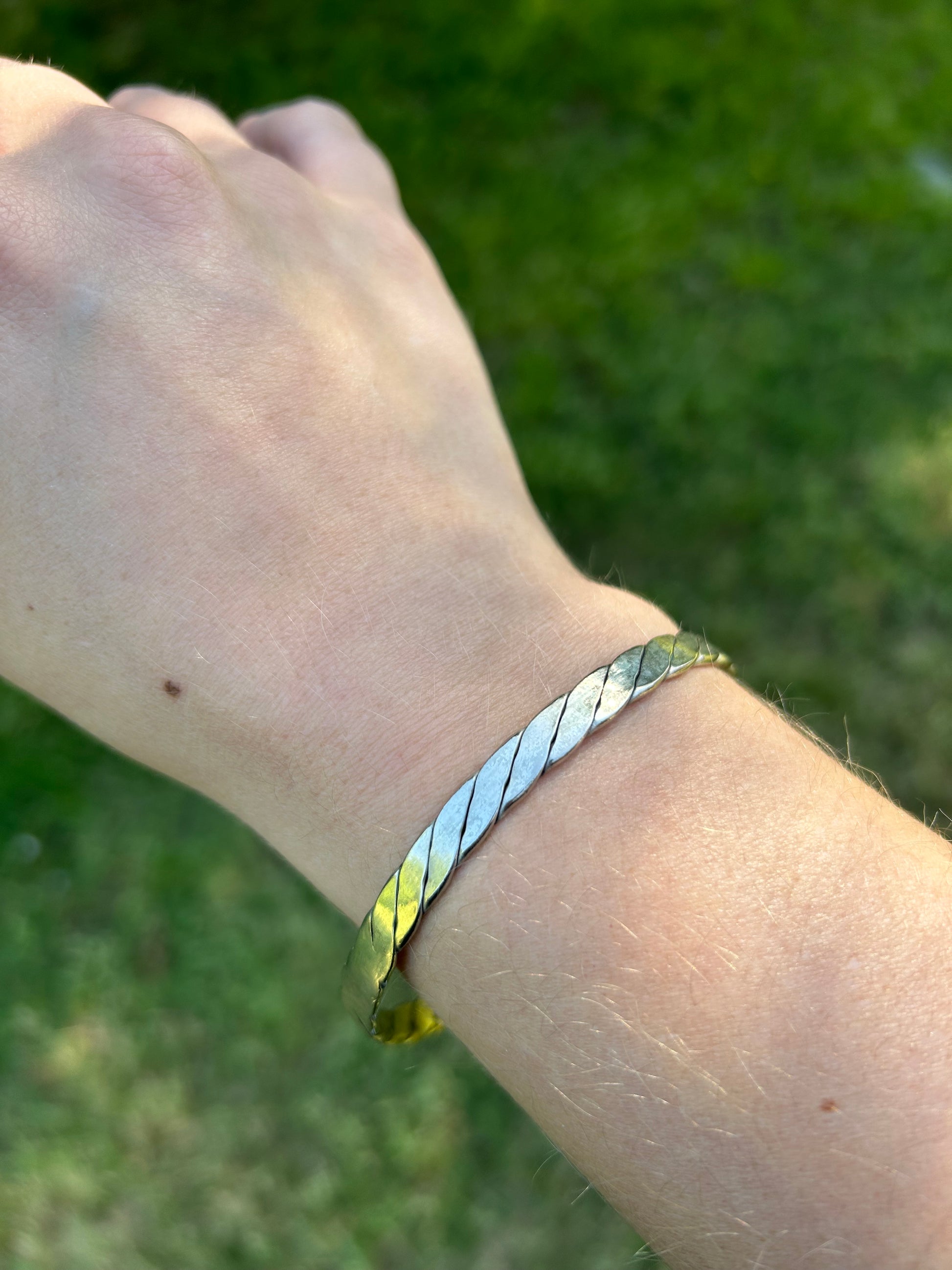 A Canyon Jewelry Lasso Bangle, a piece of western jewelry, rests on a textured burgundy tasseled rug, partially shaded by the edge of a white object, with sunlit grass visible in the background.