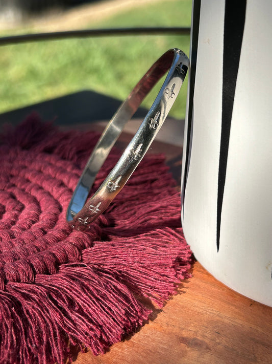 A close-up of a Canyon Jewelry Desert Charm Bangle with intricate engravings, resting on a burgundy tasseled textile next to a white mug, illuminated by natural sunlight.