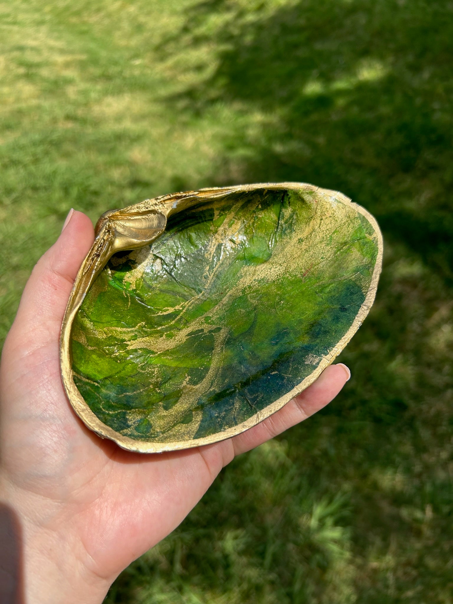 A decorative ceramic dish shaped and painted to resemble a green leaf with vein details, displayed on a sterling silver surface. The Canyon Jewelry Green Shell Trinket Tray has subtle golden accents along the edge and handle.