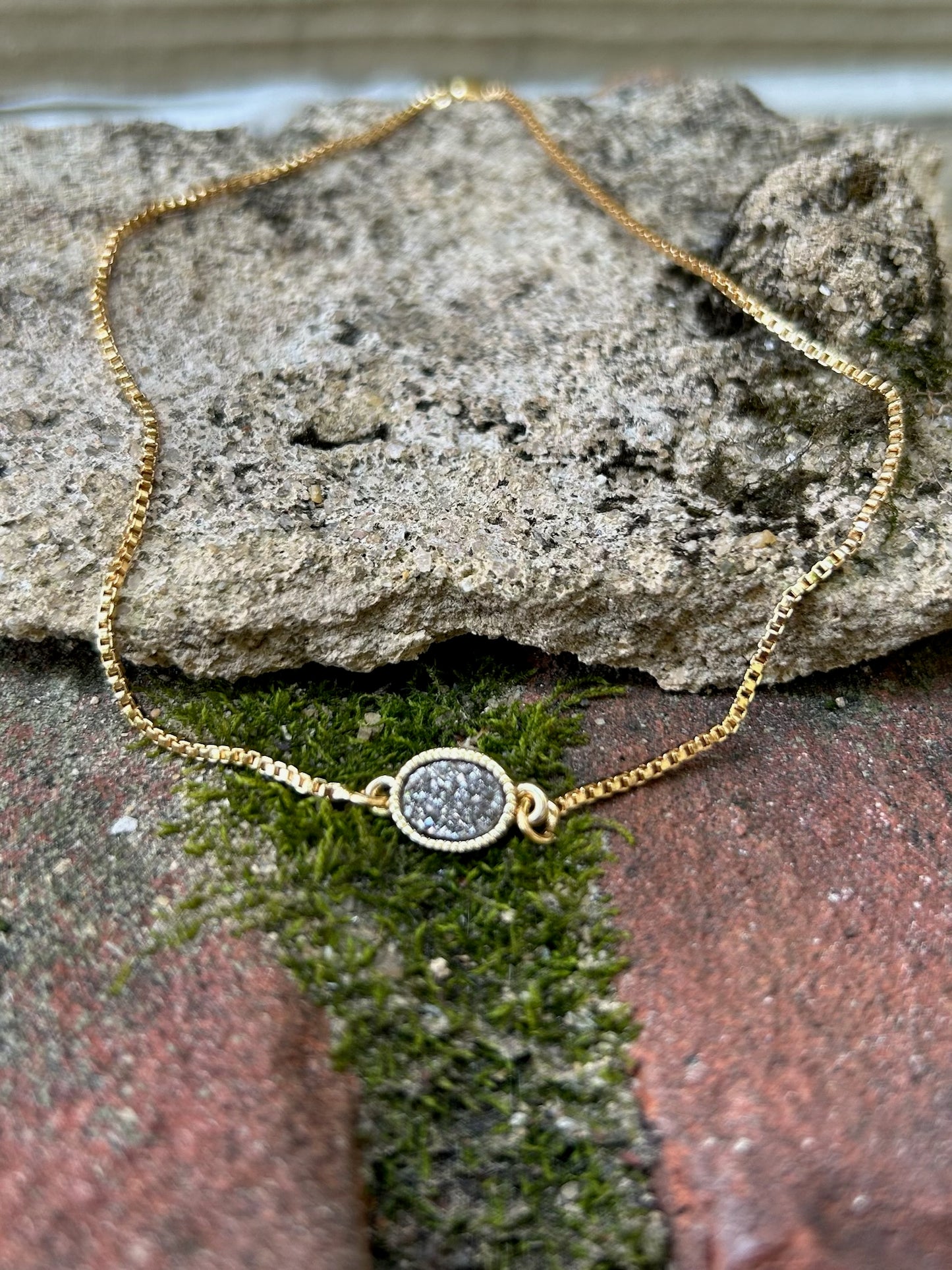 A Pedal to the Metal Bracelet with a delicate chain and a small, circular, glittering pendant lies on a wooden surface next to a burgundy fabric with fringe in western fashion.