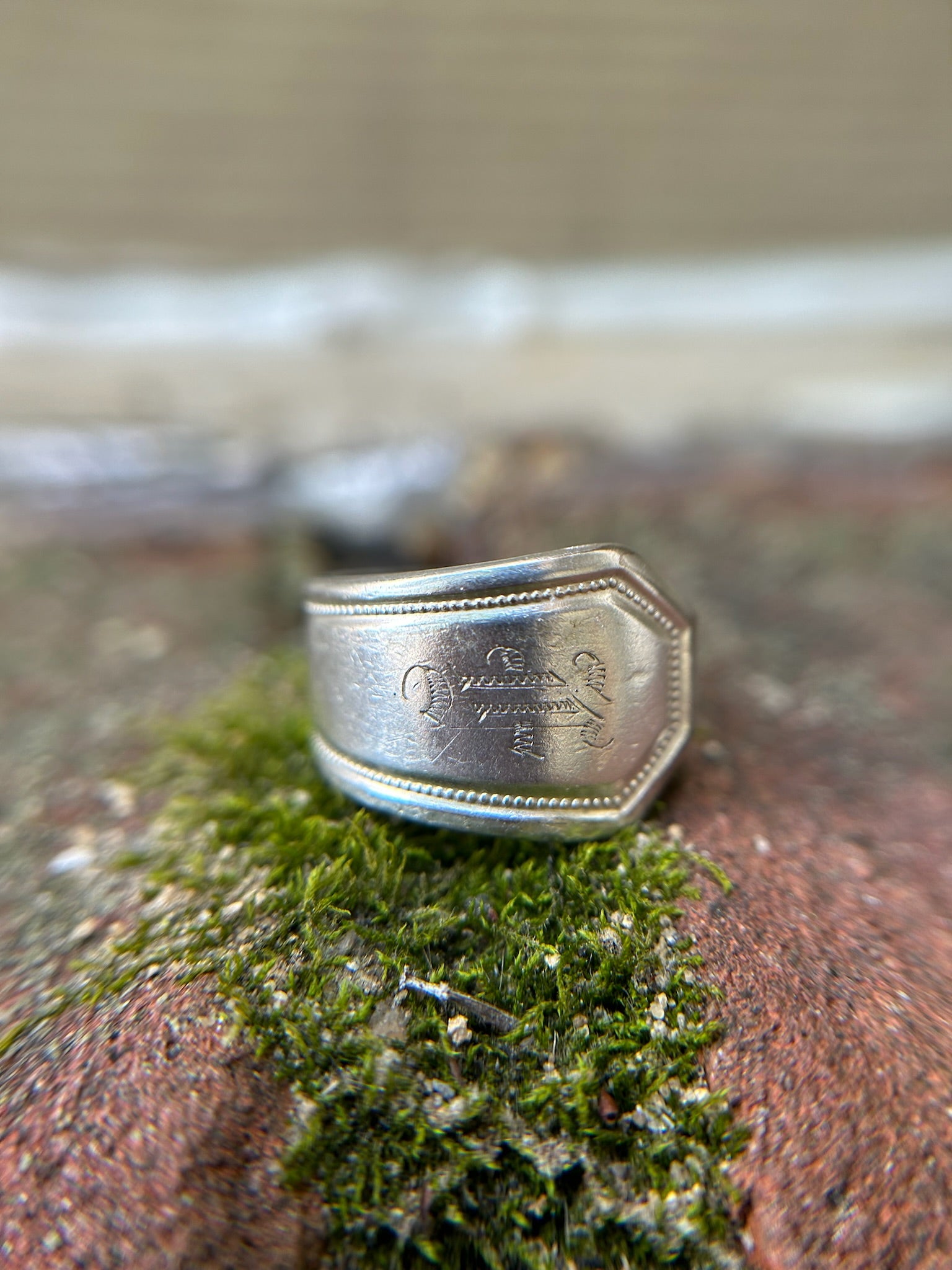 Close-up of a Vintage Rogers Spoon Ring by Canyon Jewelry partially embedded in green moss, with blurred background, focusing on the details and texture of the ring and moss.