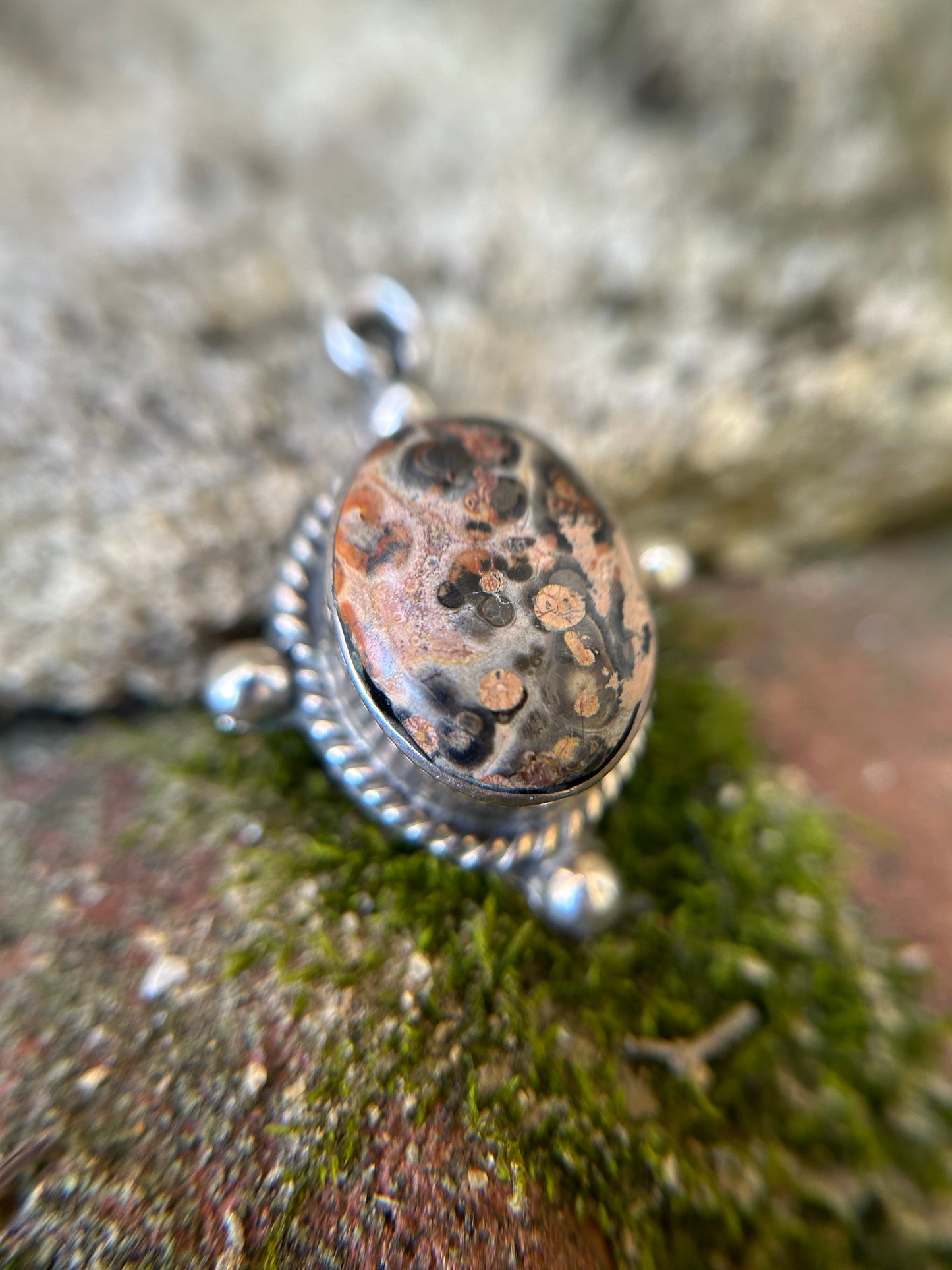 Close-up of an intricately designed Canyon Jewelry Volcano Pendant with a speckled oval stone, set in a sterling silver frame, resting on a mossy surface. The background is blurred with a focus on the detailed craftsmanship.