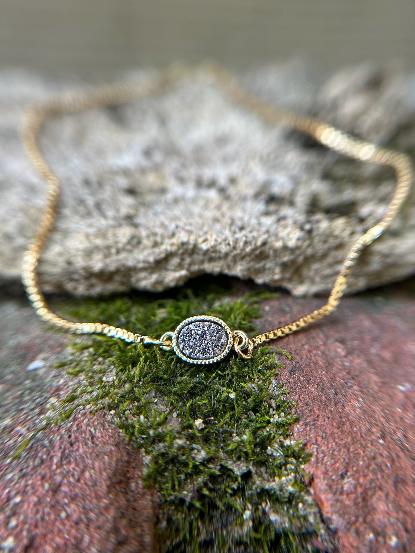 A Pedal to the Metal Bracelet with a delicate chain and a small, circular, glittering pendant lies on a wooden surface next to a burgundy fabric with fringe in western fashion.