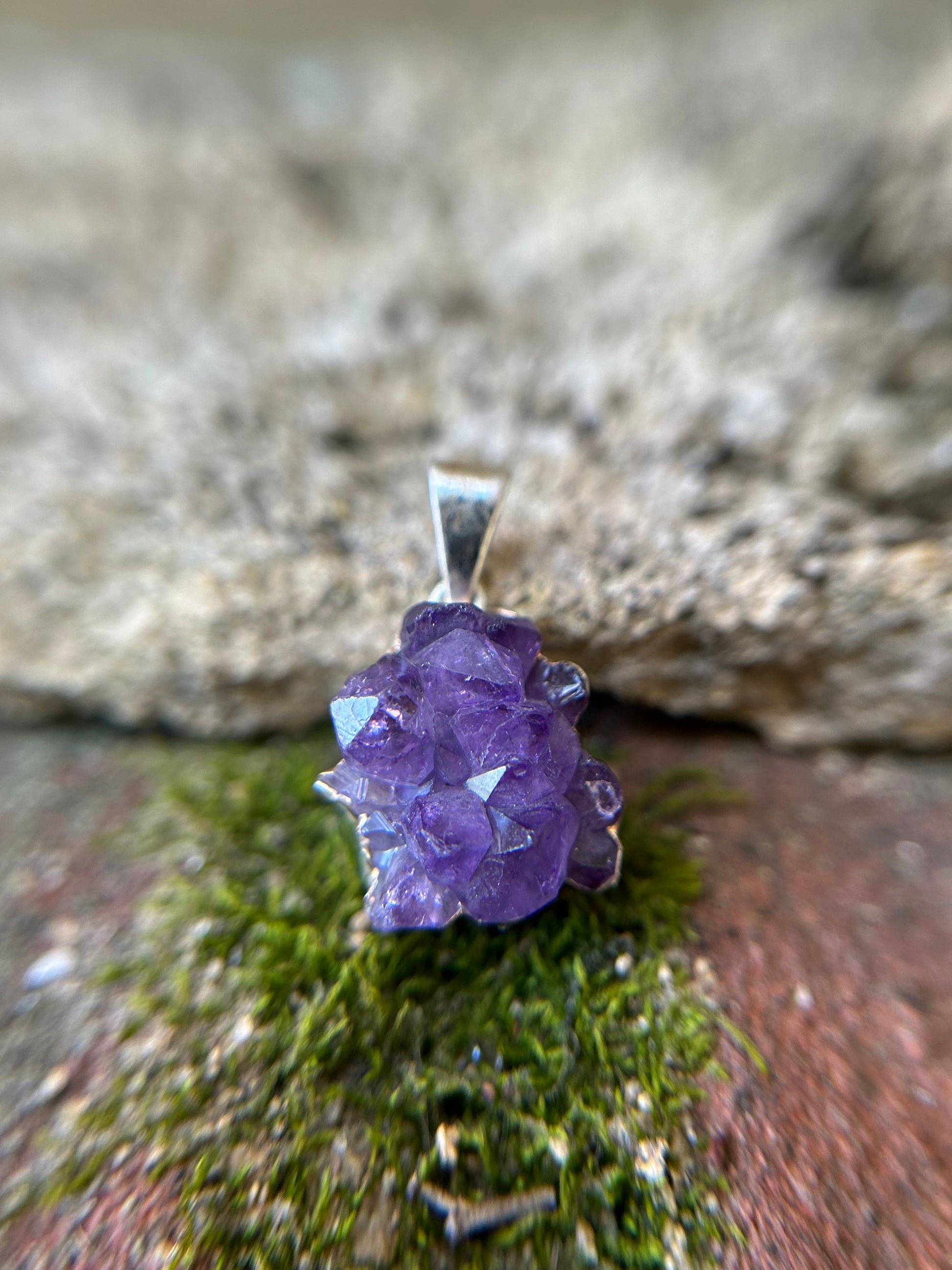 Close-up image of a purple amethyst Treasure Trove Pendant set in sterling silver by Canyon Jewelry on a background of green moss and a blurred stone surface. The crystal shows distinct facets and a vibrant hue.