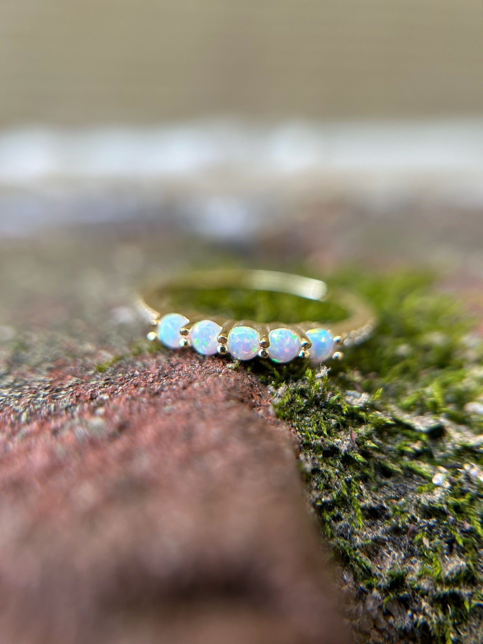 Close-up of a delicate Opalescence Ring with five opal stones, set on a moss-covered rock, showcasing Canyon Jewelry's boho jewelry style, with a blurry background emphasizing the gemstones' iridescence.