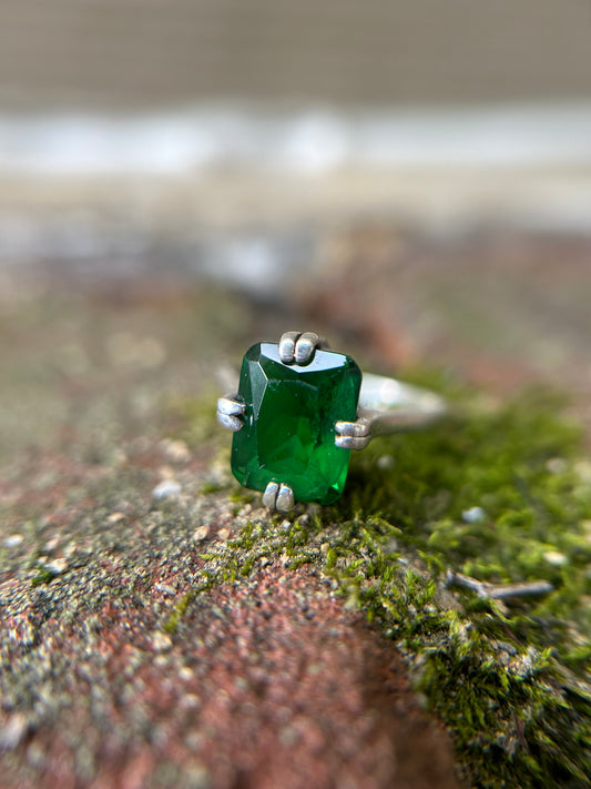 A close-up of a hand holding an Enchanted Forest Ring from Canyon Jewelry with a large, green gemstone, set against a softly blurred green grass background, capturing a vintage style.