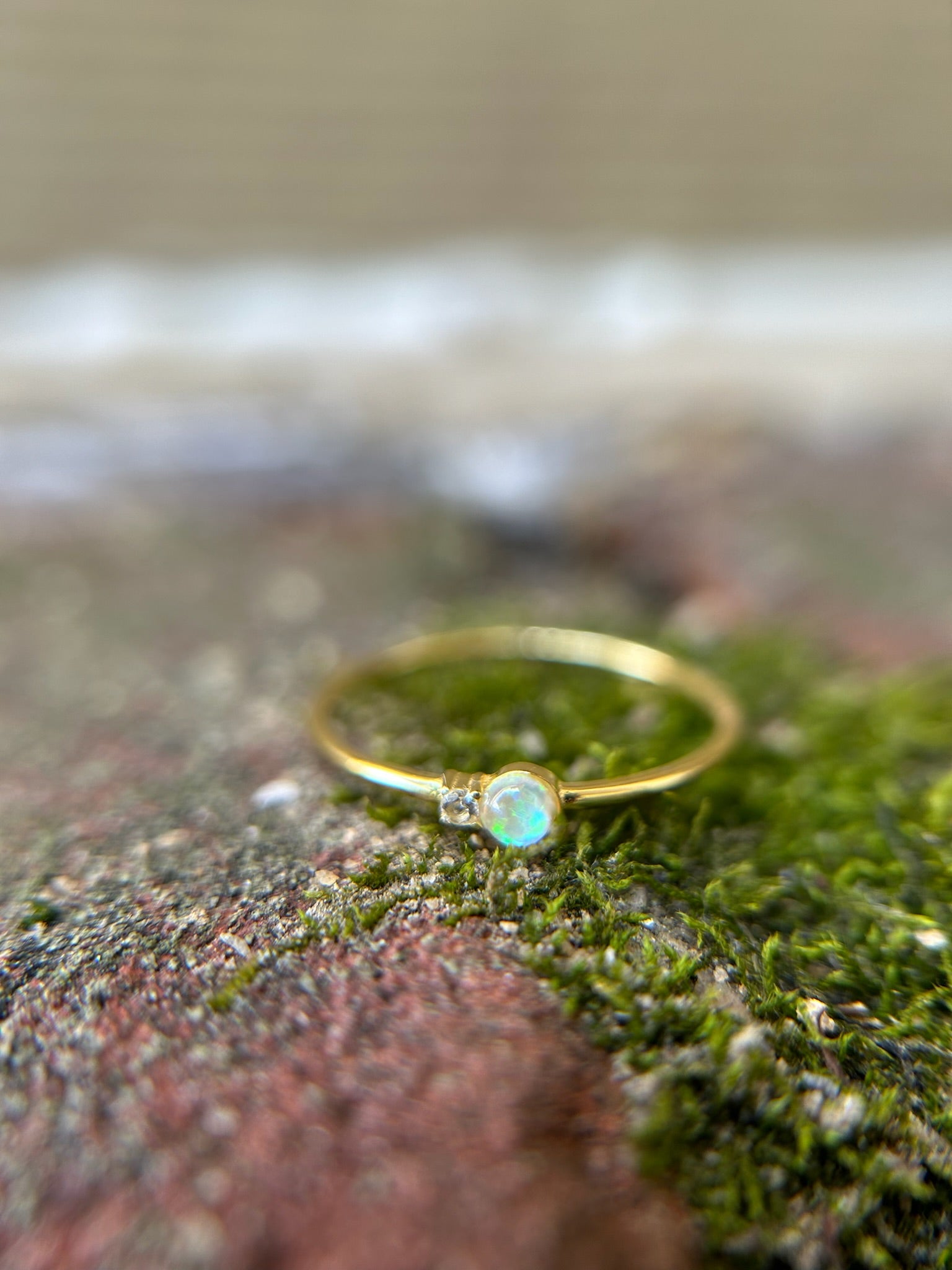 Close-up of a delicate Selene Ring by Canyon Jewelry, with a small opal set in it, resting on a textured surface covered with moss and tiny red plants. The background is softly blurred, evoking a boho vibe.