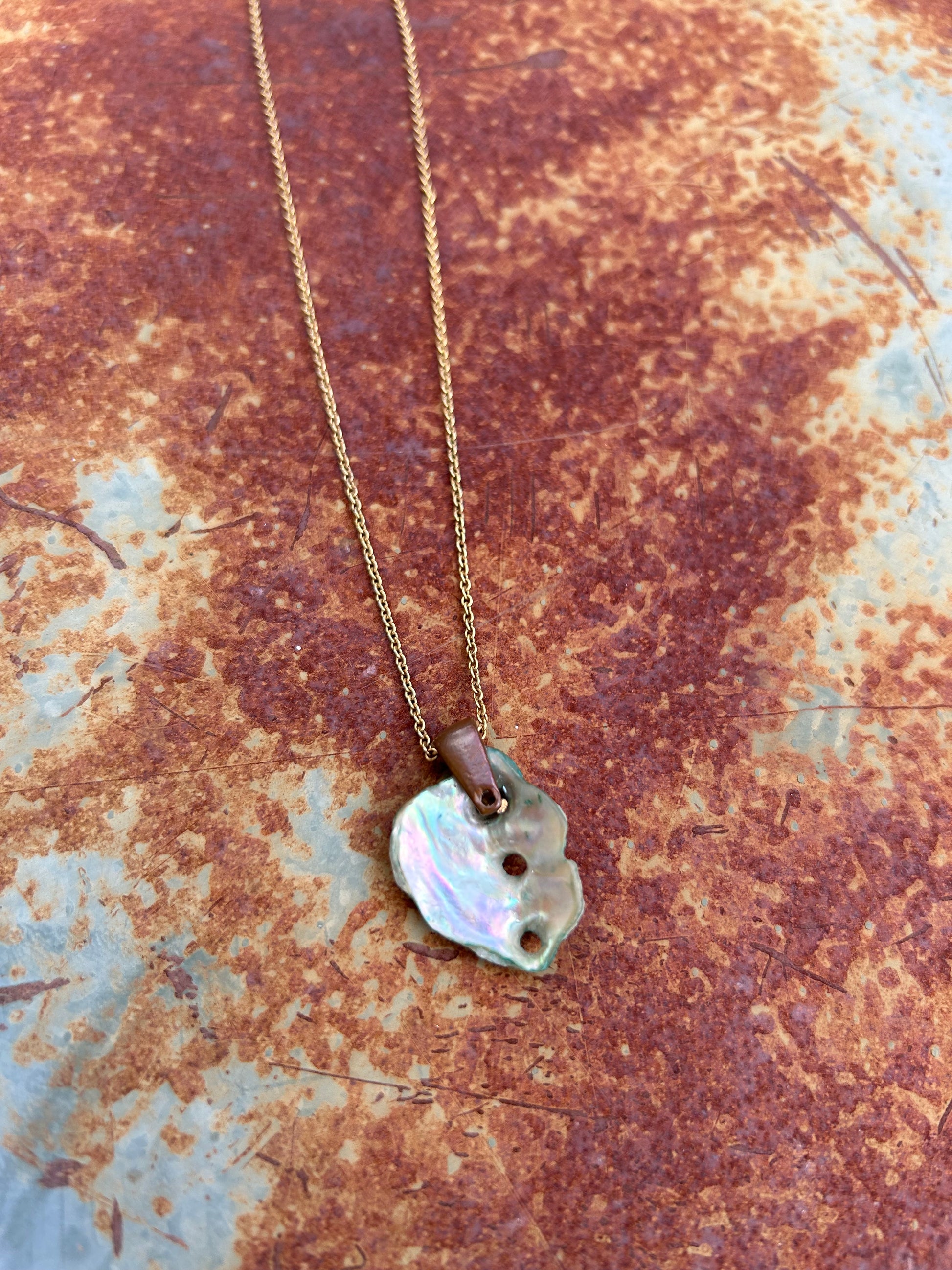 A Sandy Shores Necklace by Canyon Jewelry featuring a pearlescent shell pendant on a gold chain necklace rests on a wooden surface beside a black and white striped vase and a green patterned pot.