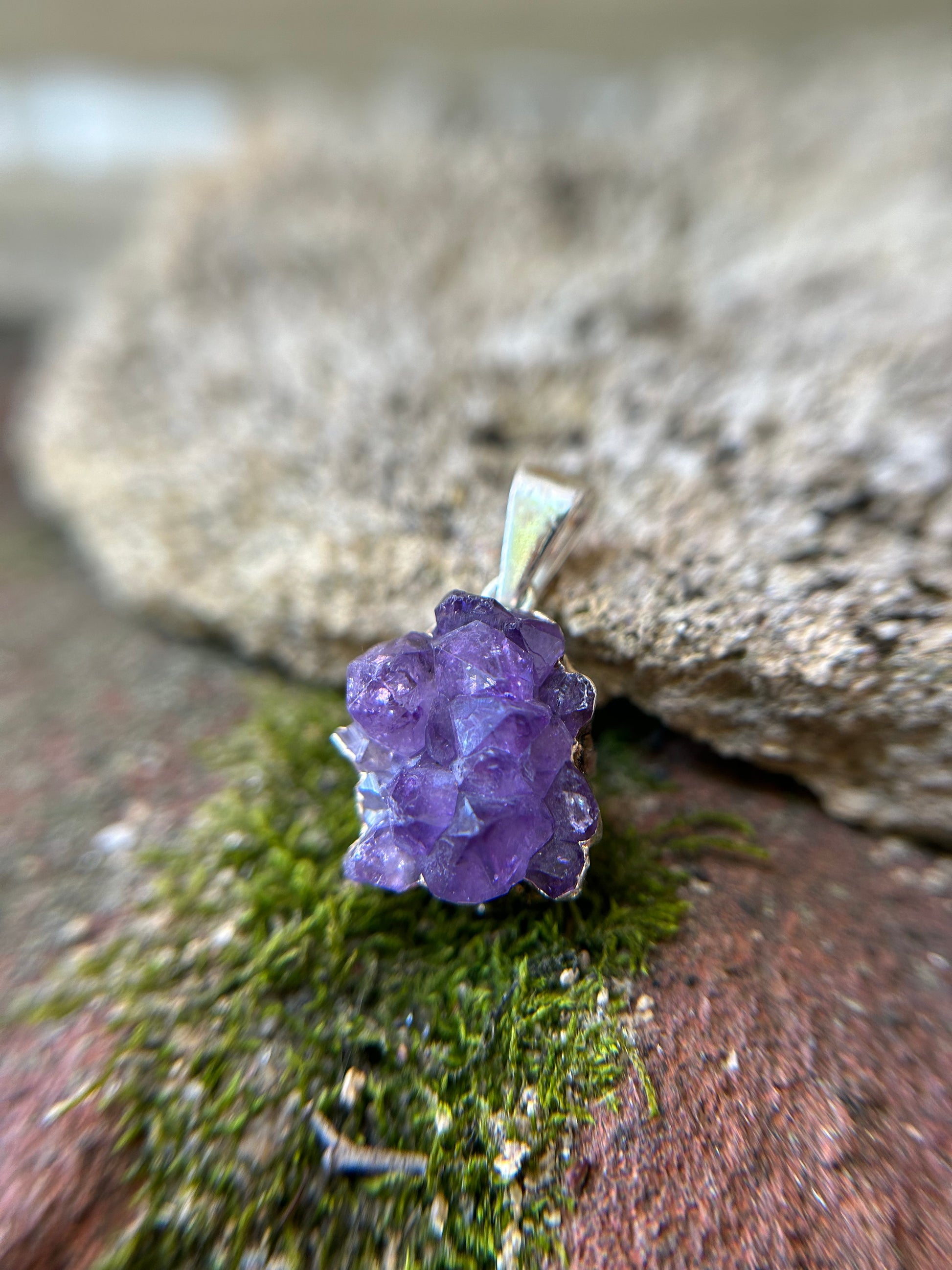 Close-up image of a purple amethyst Treasure Trove Pendant set in sterling silver by Canyon Jewelry on a background of green moss and a blurred stone surface. The crystal shows distinct facets and a vibrant hue.