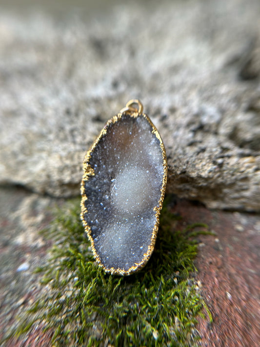 Close-up of a translucent, oval Druzy Agate pendant with a sparkling, crystalline interior and a sterling silver rim, resting on a moss-covered rock, with a blurred background. (Brand Name: Canyon Jewelry)