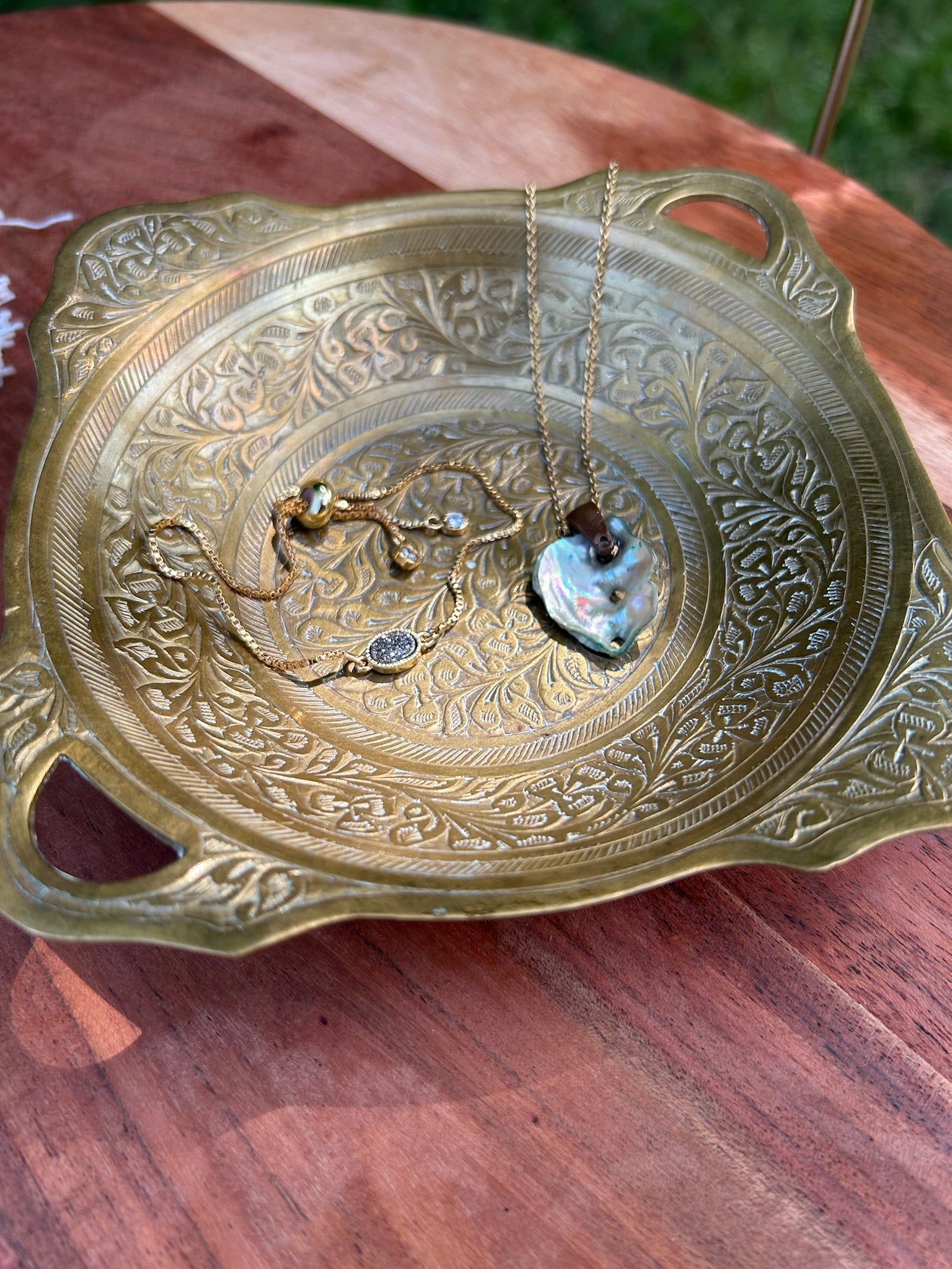 An intricately designed Canyon Jewelry Brass Trinket Tray with boho jewelry patterns placed on a wooden surface outdoors, partially shaded by sunlight.