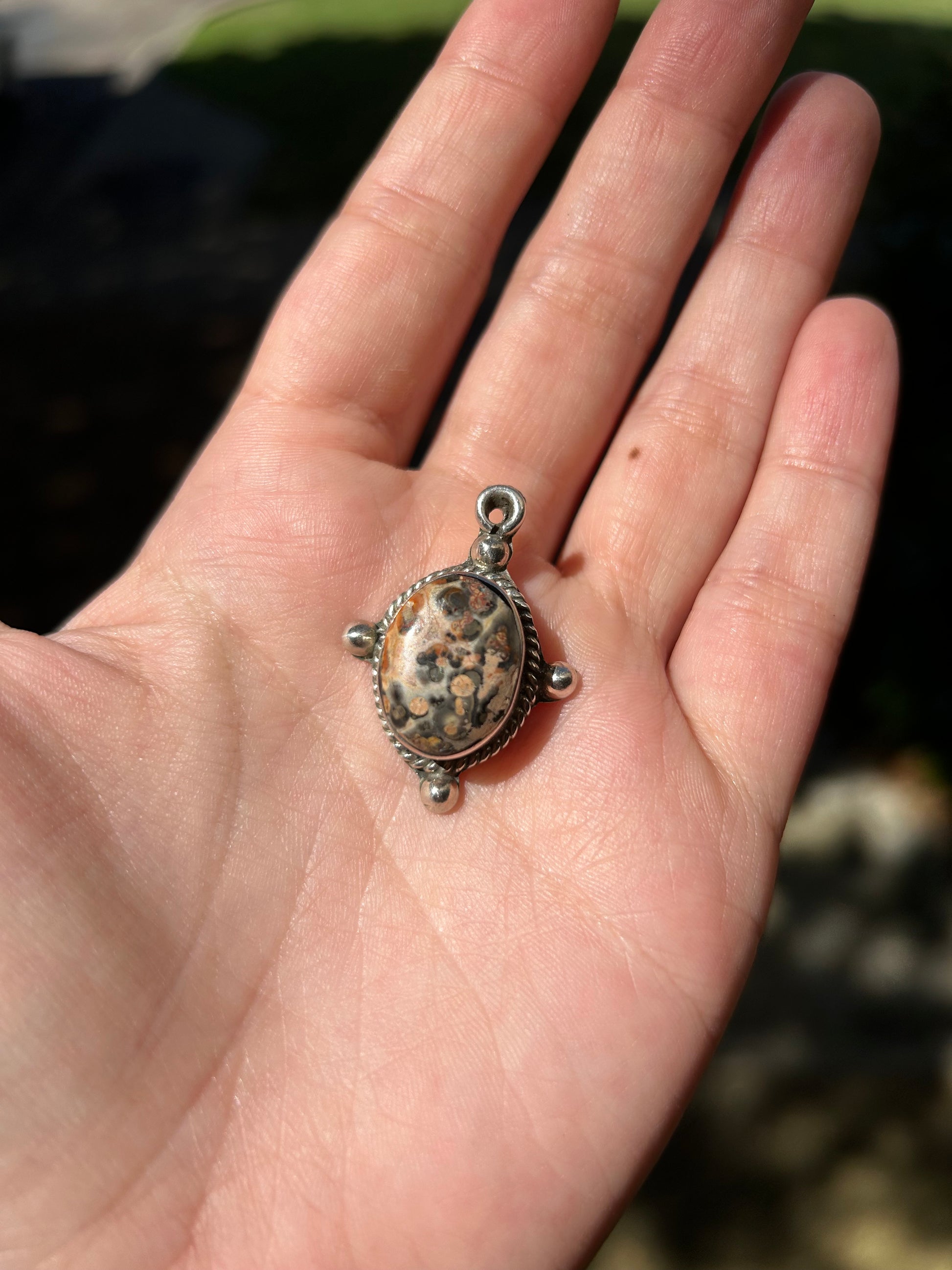 Close-up of an intricately designed Canyon Jewelry Volcano Pendant with a speckled oval stone, set in a sterling silver frame, resting on a mossy surface. The background is blurred with a focus on the detailed craftsmanship.