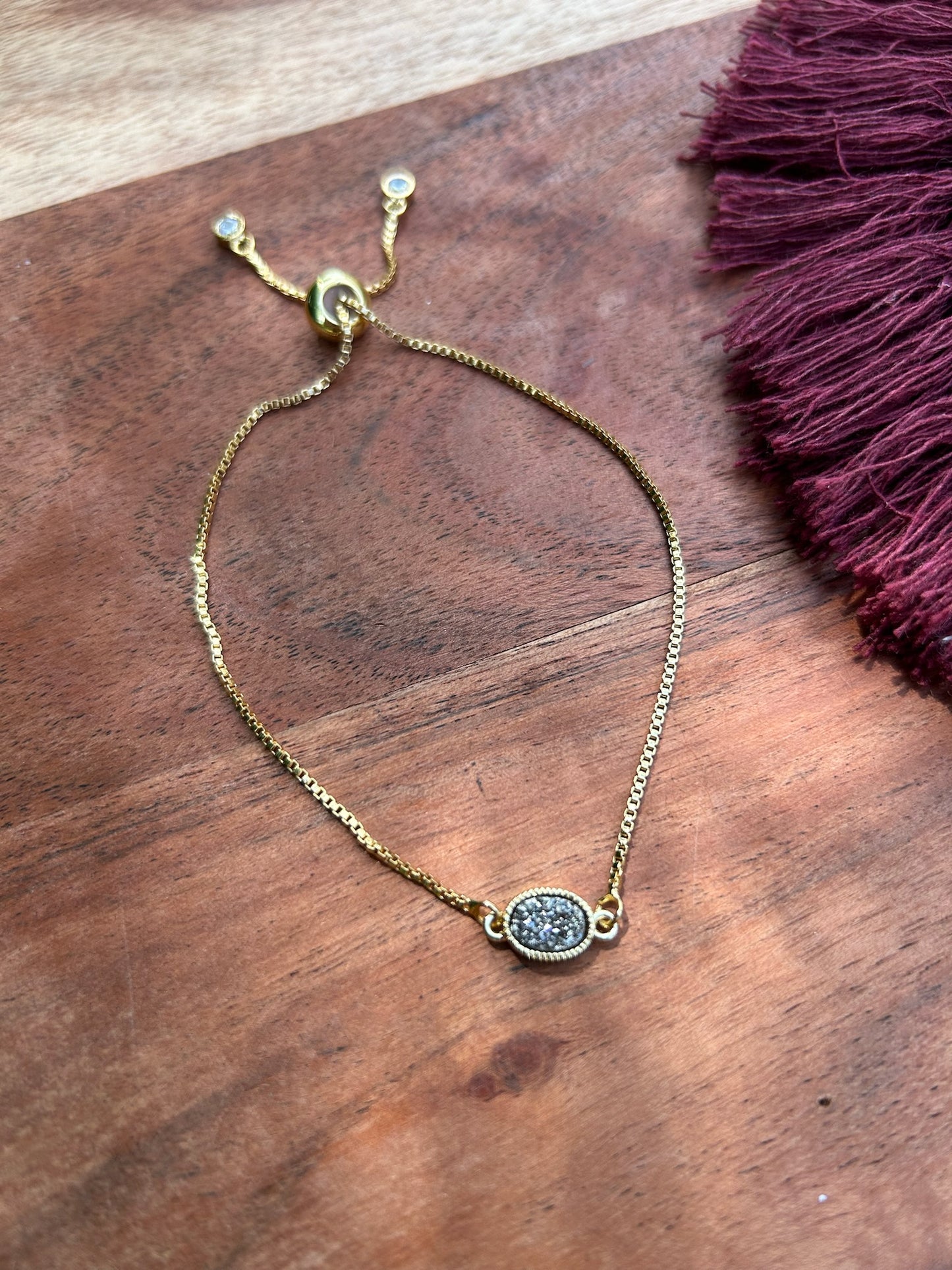 A Pedal to the Metal Bracelet with a delicate chain and a small, circular, glittering pendant lies on a wooden surface next to a burgundy fabric with fringe in western fashion.