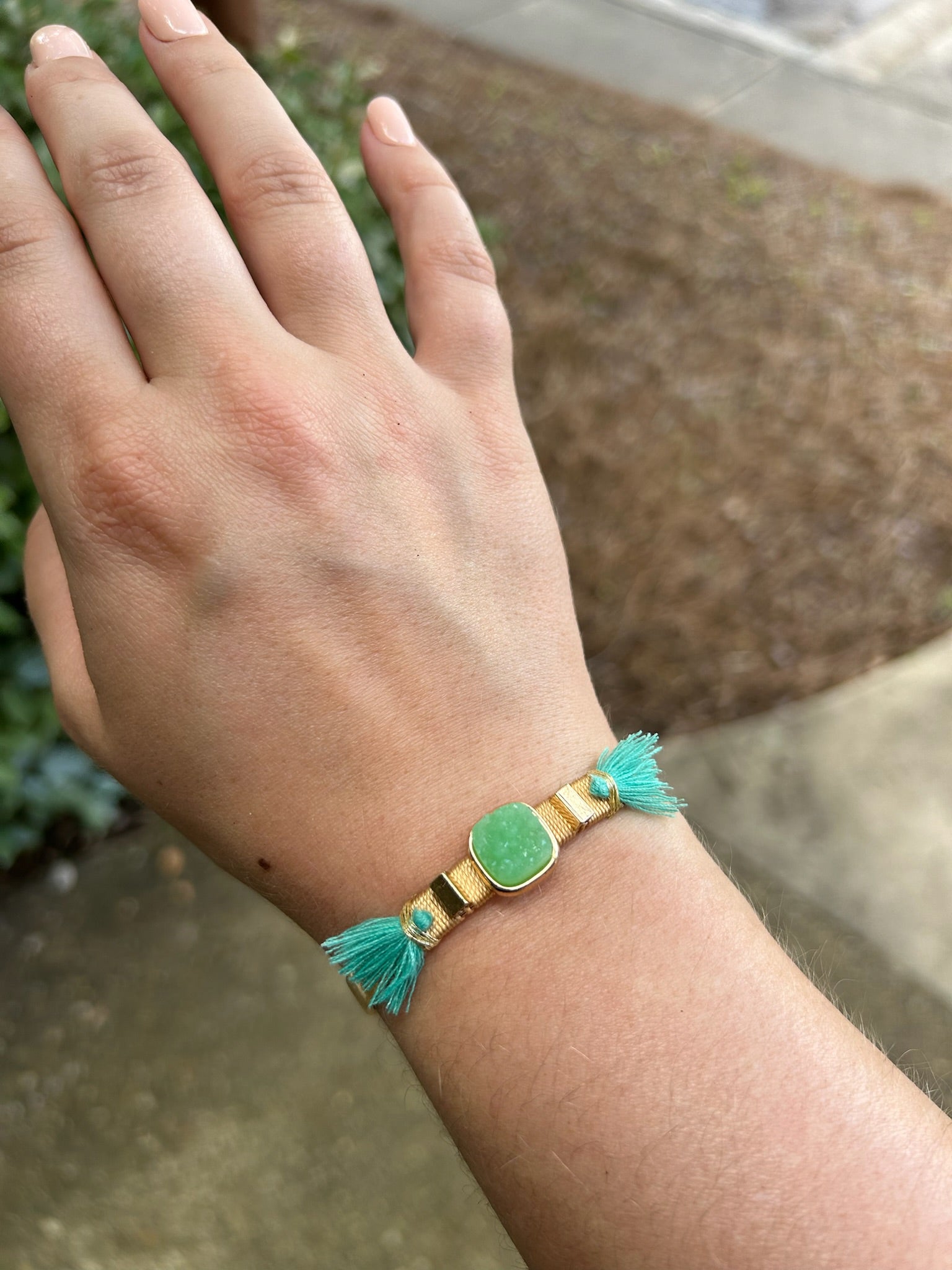 A person's wrist adorned with a stylish Painted Pony Bracelet from Canyon Jewelry featuring a large green oval centerpiece and turquoise tassel details, set against a soft-focus outdoor background. This piece exemplifies statement jewelry.