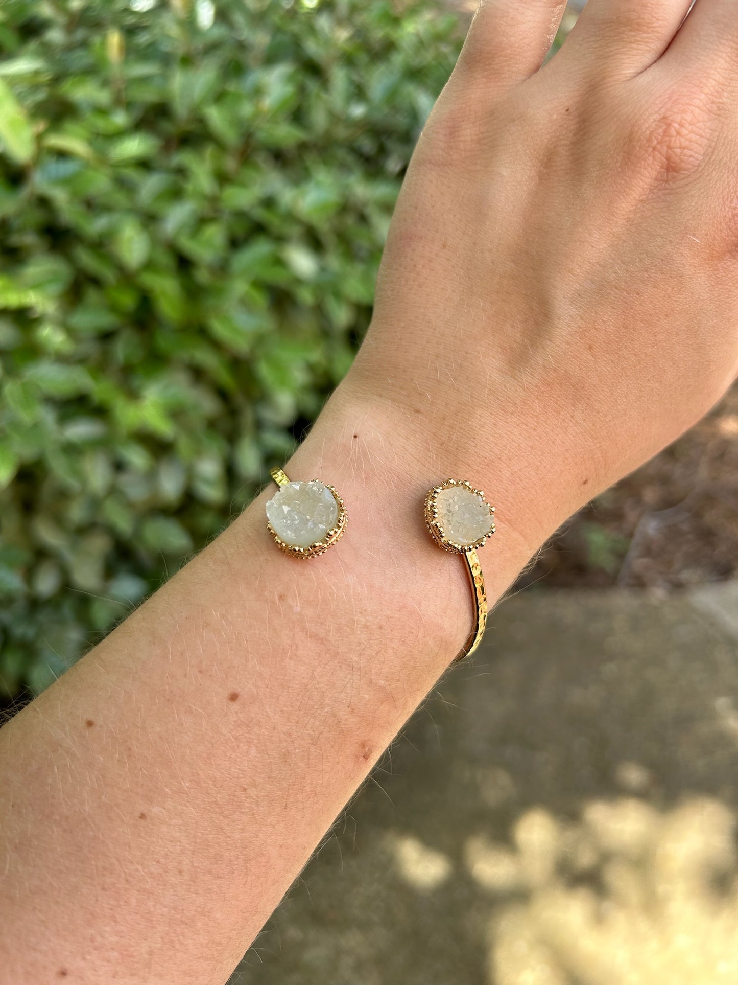 A close-up of a hand holding two Divergence Bracelets from Canyon Jewelry, with large, white, sparkly circular pendants, focuses against a softly blurred greenery background. These statement jewelry pieces capture attention effortlessly.