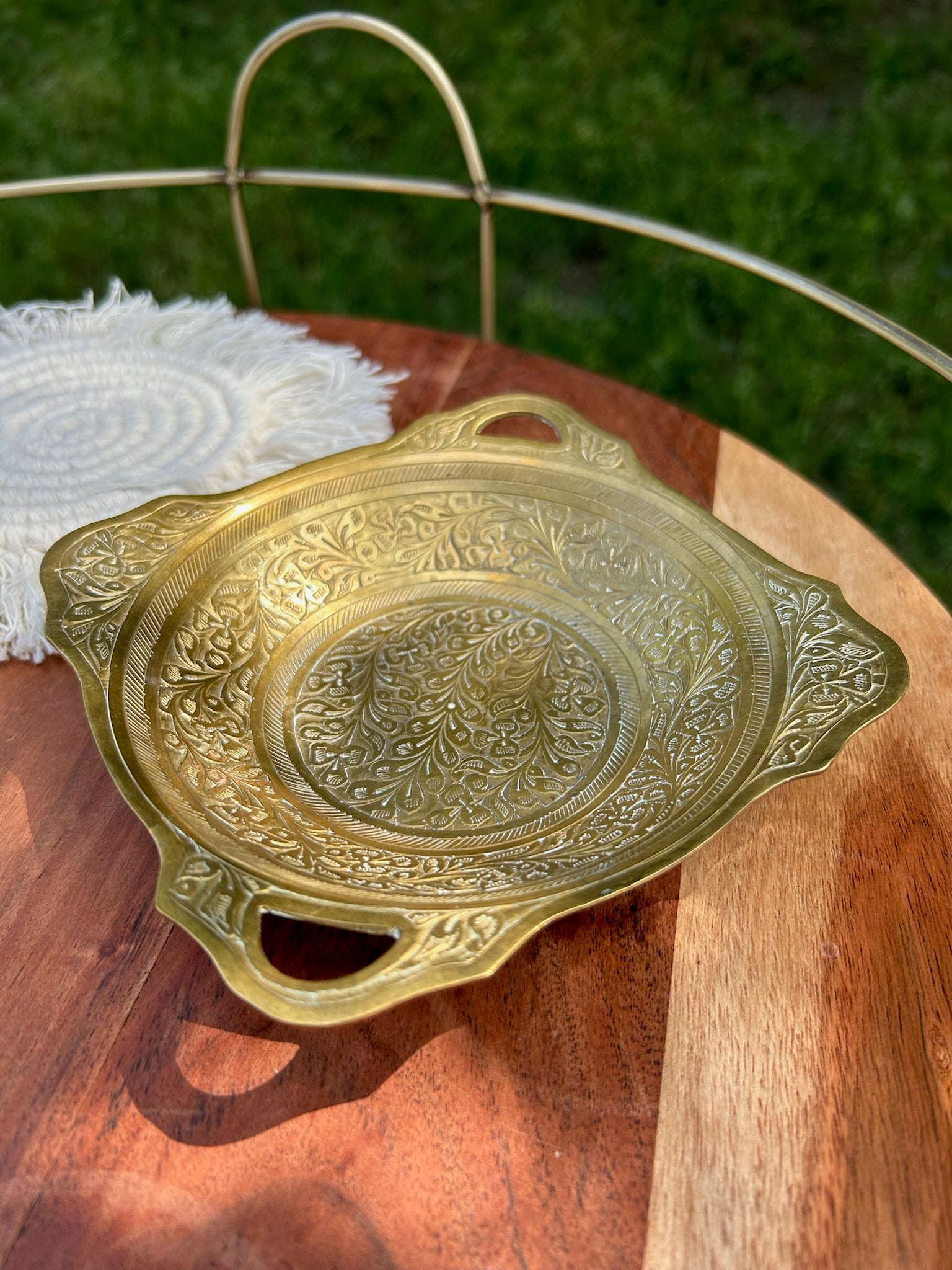 An intricately designed Canyon Jewelry Brass Trinket Tray with boho jewelry patterns placed on a wooden surface outdoors, partially shaded by sunlight.