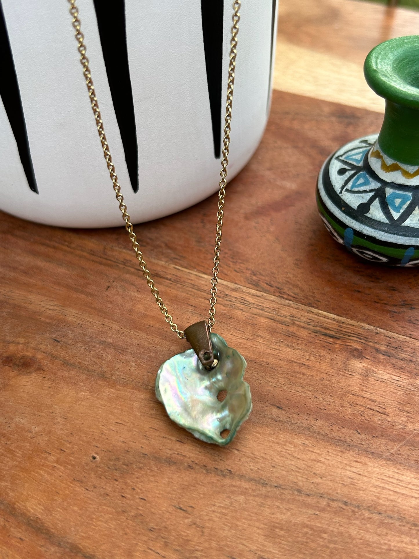 A Sandy Shores Necklace by Canyon Jewelry featuring a pearlescent shell pendant on a gold chain necklace rests on a wooden surface beside a black and white striped vase and a green patterned pot.