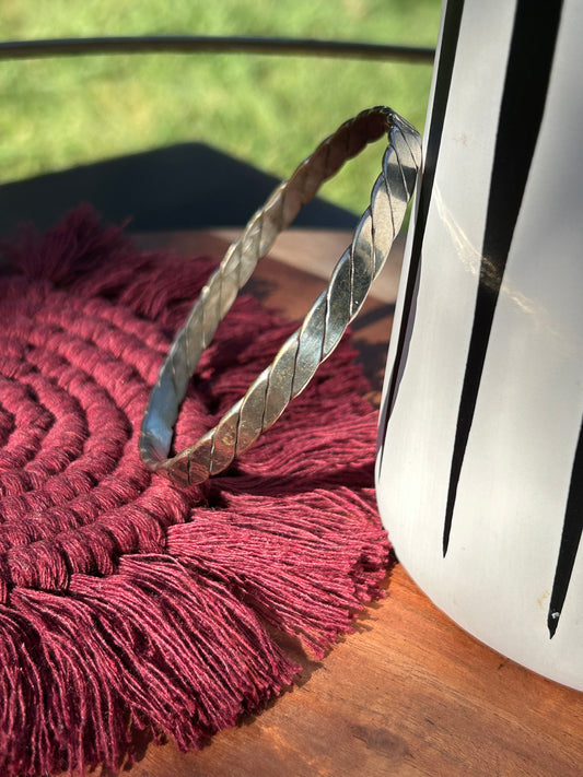 A Canyon Jewelry Lasso Bangle, a piece of western jewelry, rests on a textured burgundy tasseled rug, partially shaded by the edge of a white object, with sunlit grass visible in the background.