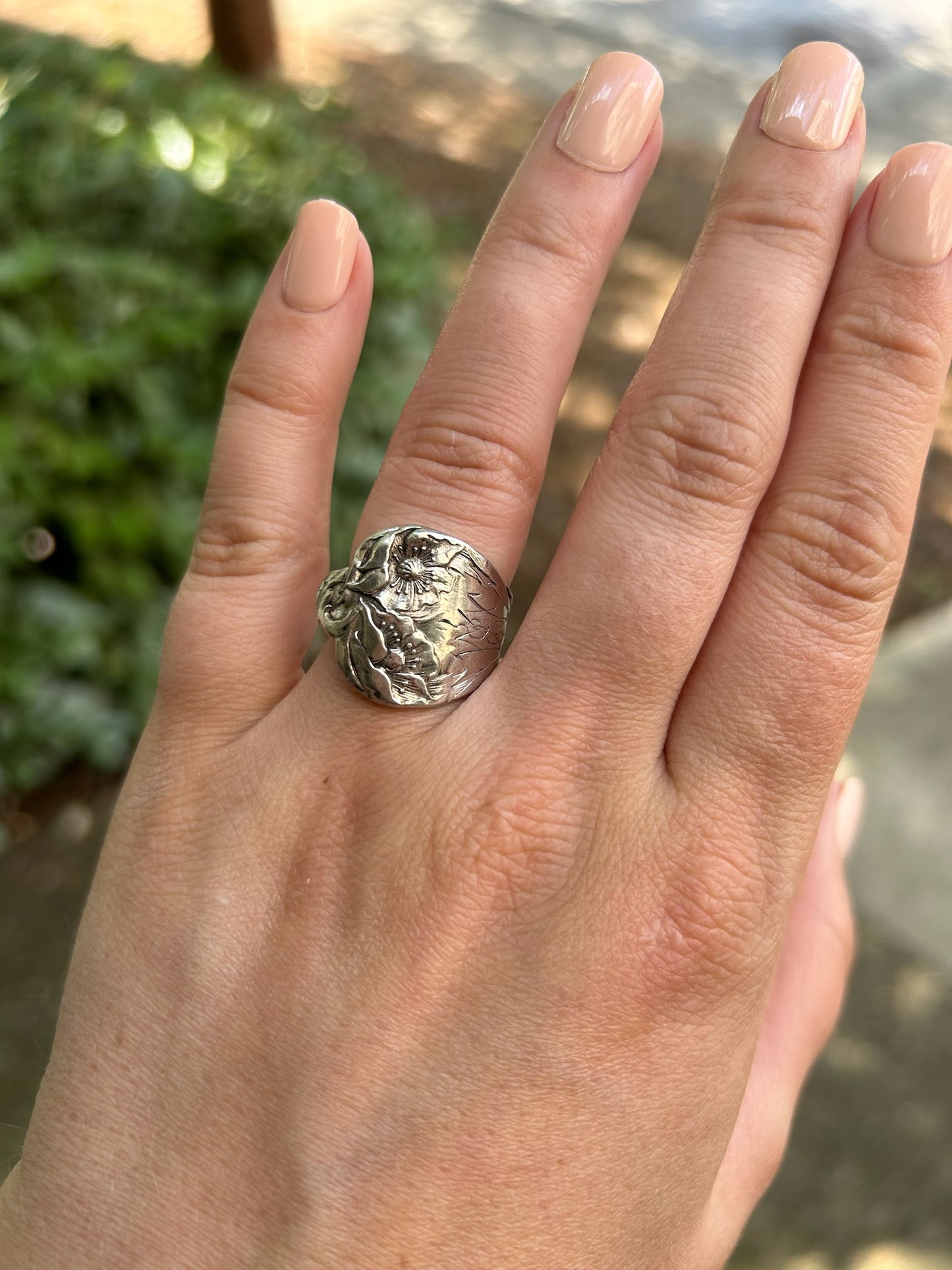 A close-up of a Canyon Jewelry 1913 Spoon Ring resting on a green mossy surface, depicting floral designs with prominent details against a blurred background.