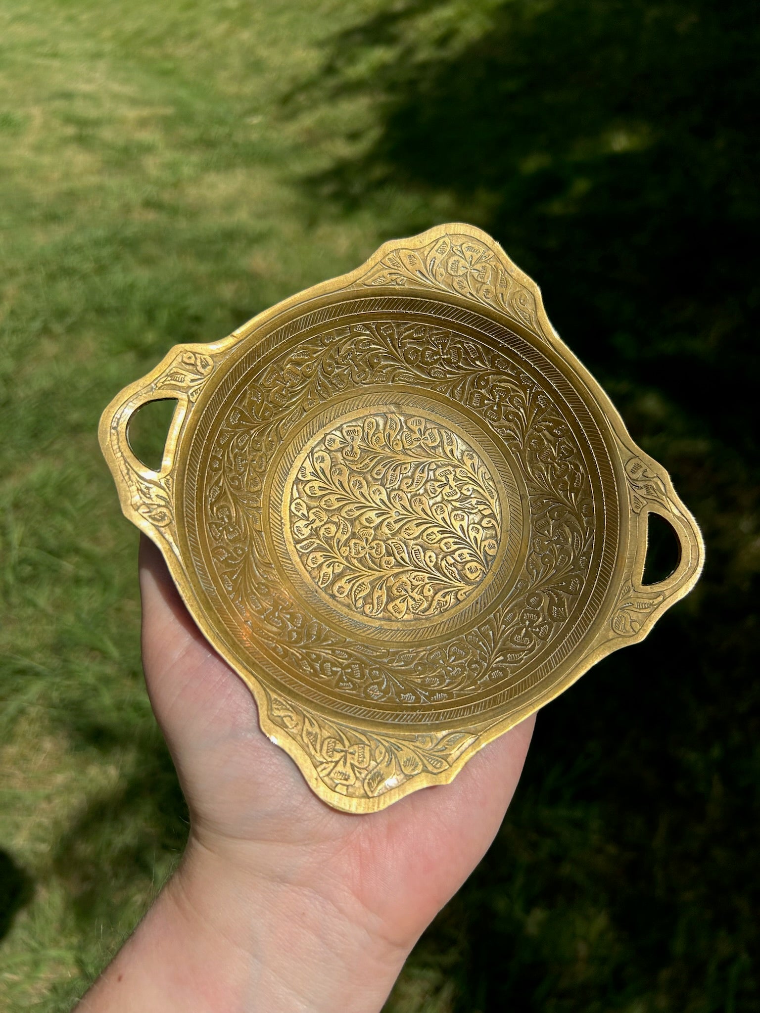 An intricately designed Canyon Jewelry Brass Trinket Tray with boho jewelry patterns placed on a wooden surface outdoors, partially shaded by sunlight.