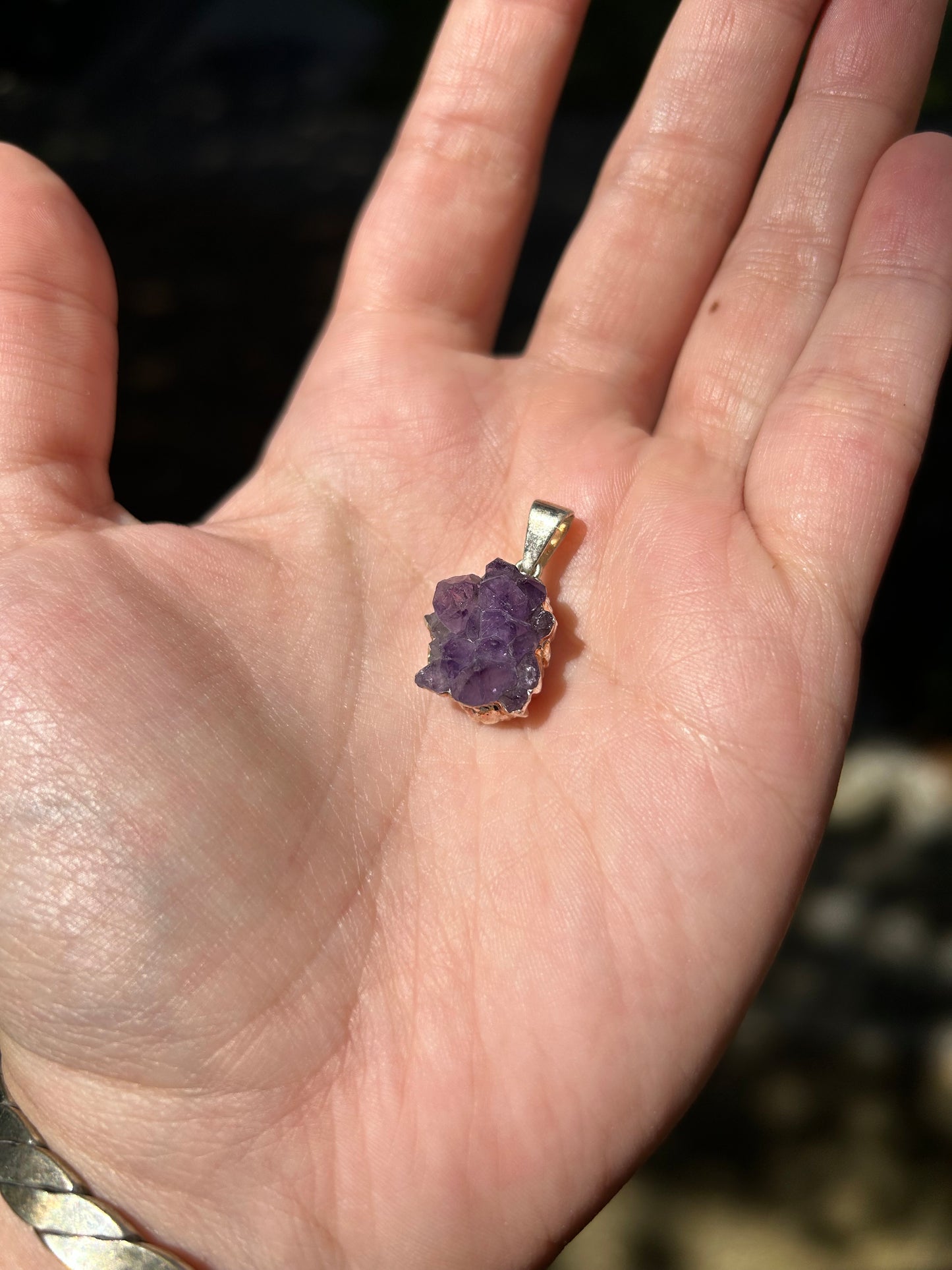 Close-up image of a purple amethyst Treasure Trove Pendant set in sterling silver by Canyon Jewelry on a background of green moss and a blurred stone surface. The crystal shows distinct facets and a vibrant hue.