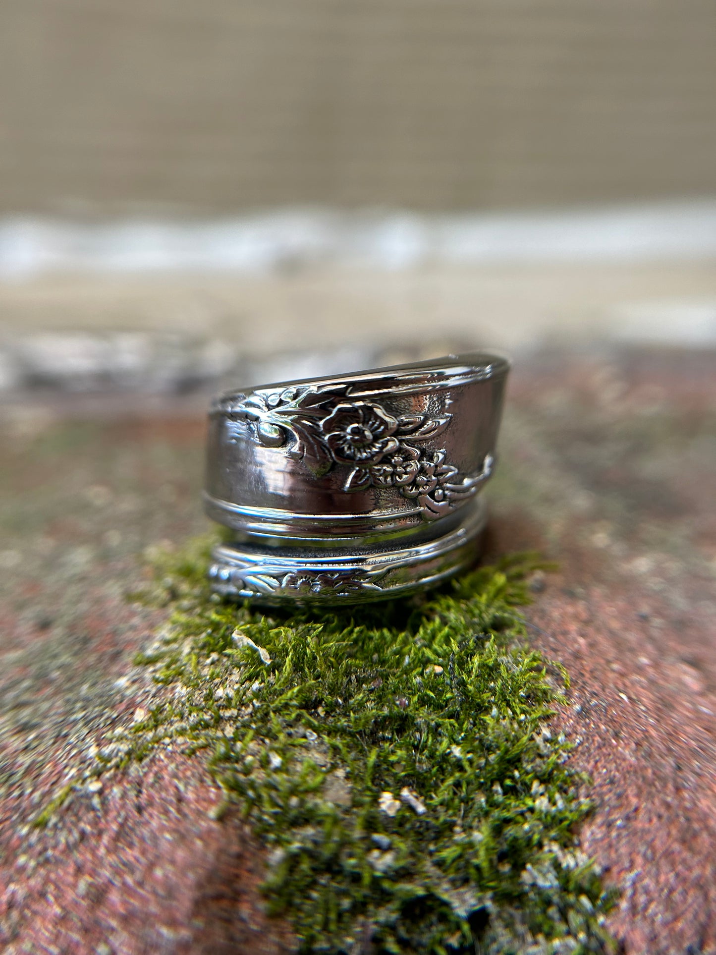 A close-up photograph of a detailed Nefertiti Spoon Ring by Canyon Jewelry with floral patterns, resting on a bed of vibrant green moss.