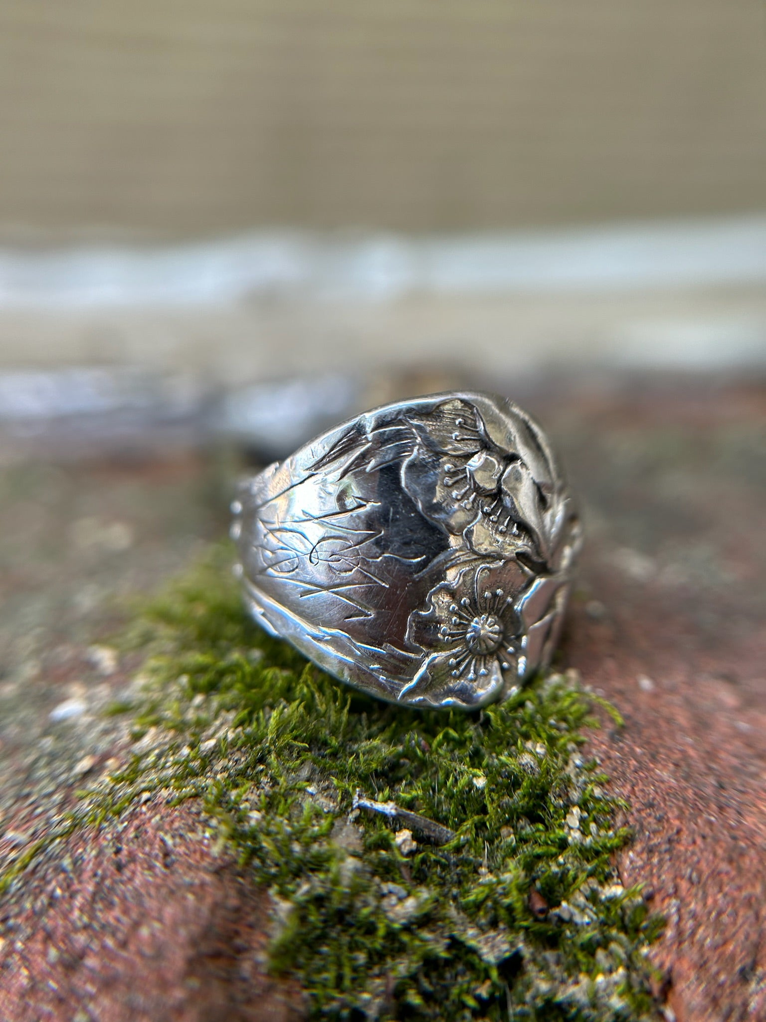 A close-up of a Canyon Jewelry 1913 Spoon Ring resting on a green mossy surface, depicting floral designs with prominent details against a blurred background.