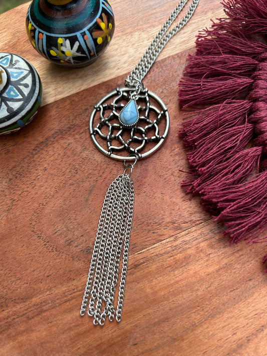 A Canyon Jewelry Dreamcatcher Necklace with a blue stone in the center and chain tassels, lying on a wooden surface next to colorful decorated pots and a red tassel.