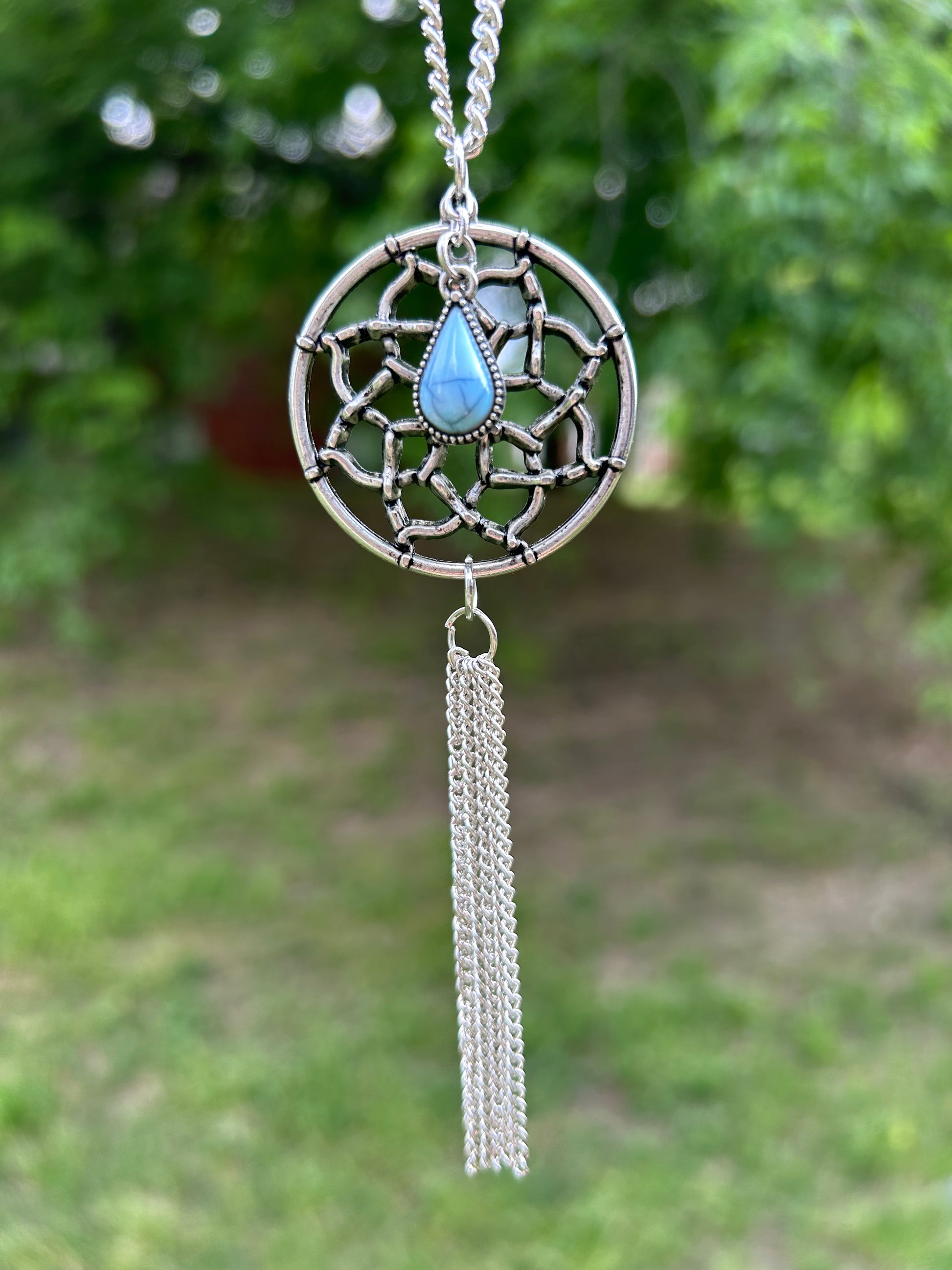 A Canyon Jewelry Dreamcatcher Necklace with a blue stone in the center and chain tassels, lying on a wooden surface next to colorful decorated pots and a red tassel.