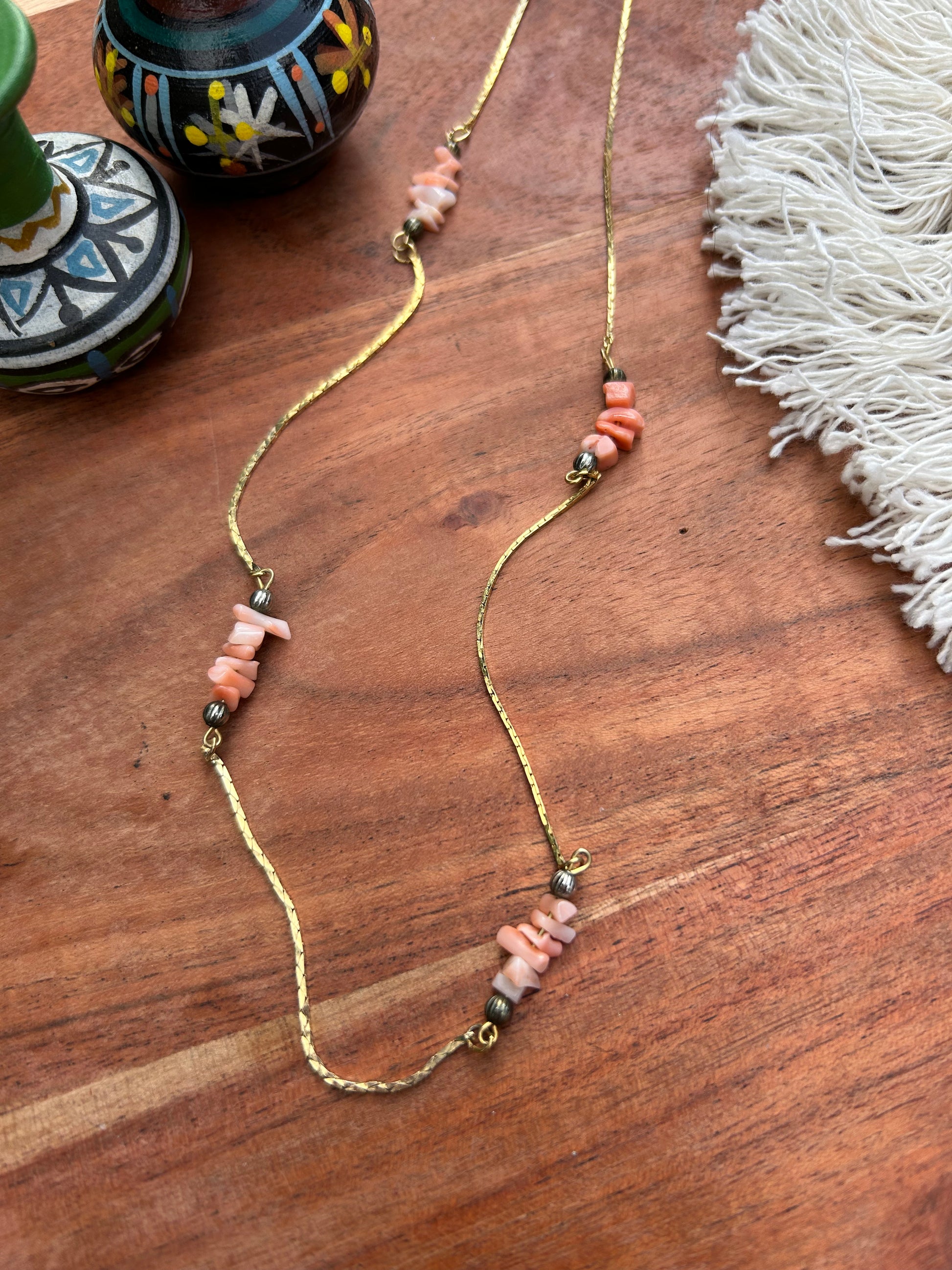 A delicate Canyon Jewelry sterling silver Pink Lady Necklace with pink and coral beads, spaced with small silver beads, laid out on a wooden surface next to decorative pottery and a fringe of a white textile.