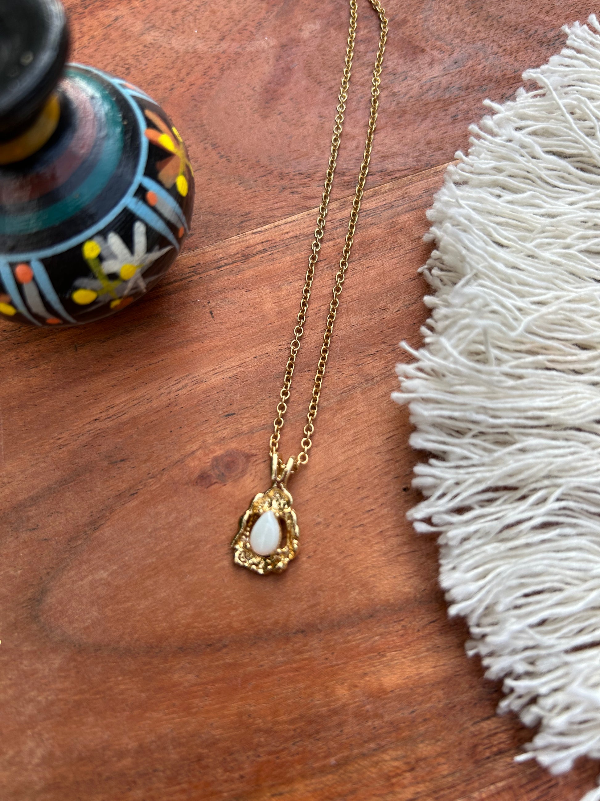 A delicate Canyon Jewelry gold Teardrop Necklace with a white oval pendant rests on a wooden surface beside a colorful boho ceramic vase and a white fringed textile.