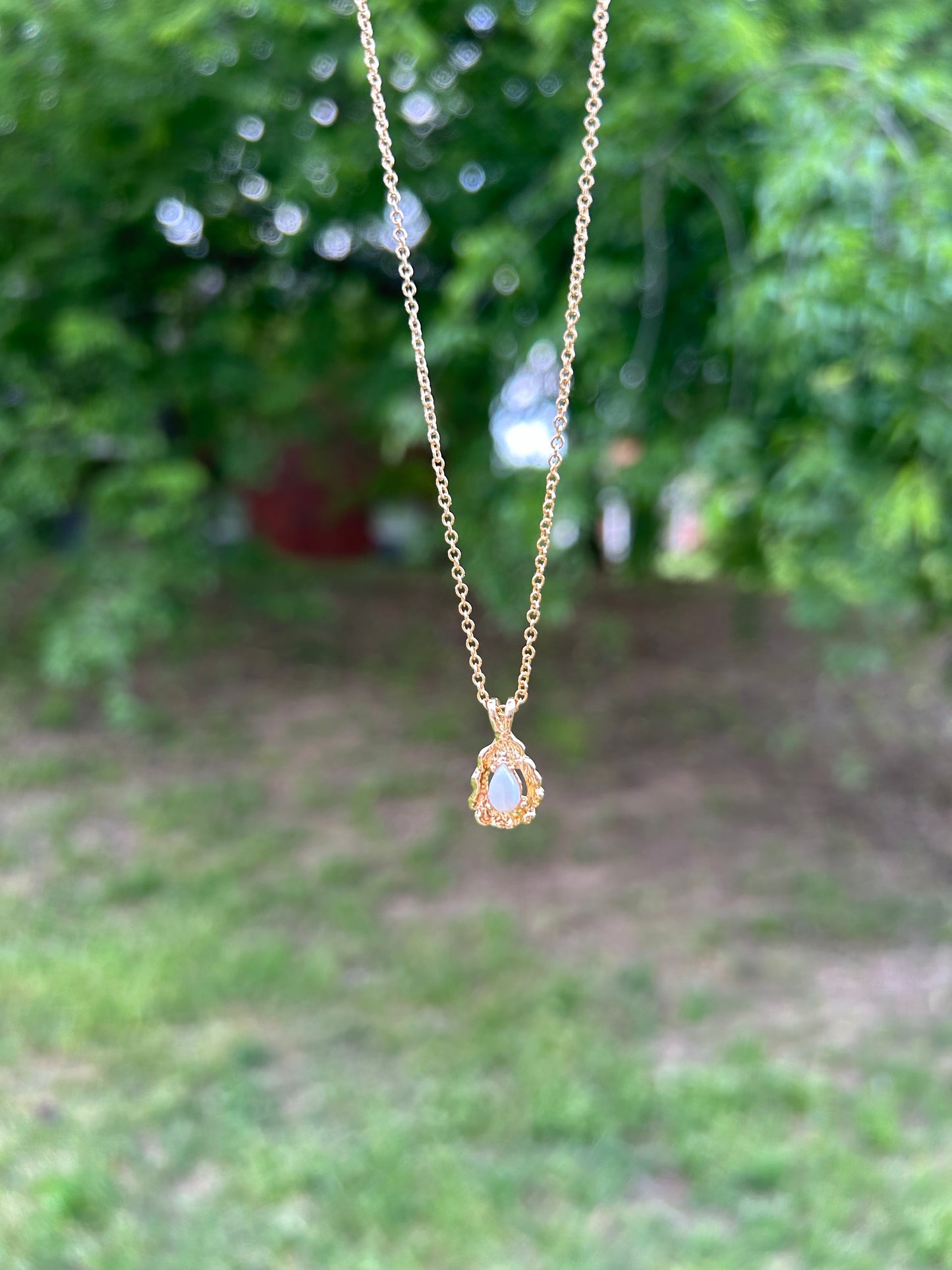 A delicate Canyon Jewelry gold Teardrop Necklace with a white oval pendant rests on a wooden surface beside a colorful boho ceramic vase and a white fringed textile.