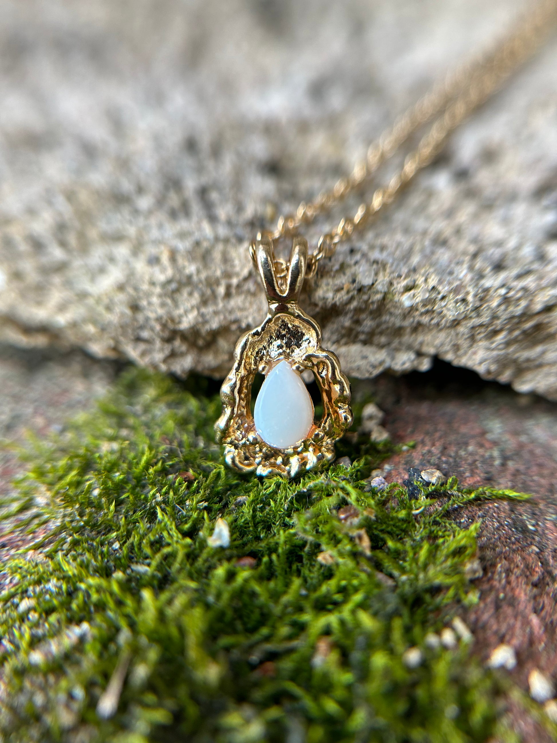 A delicate Canyon Jewelry gold Teardrop Necklace with a white oval pendant rests on a wooden surface beside a colorful boho ceramic vase and a white fringed textile.